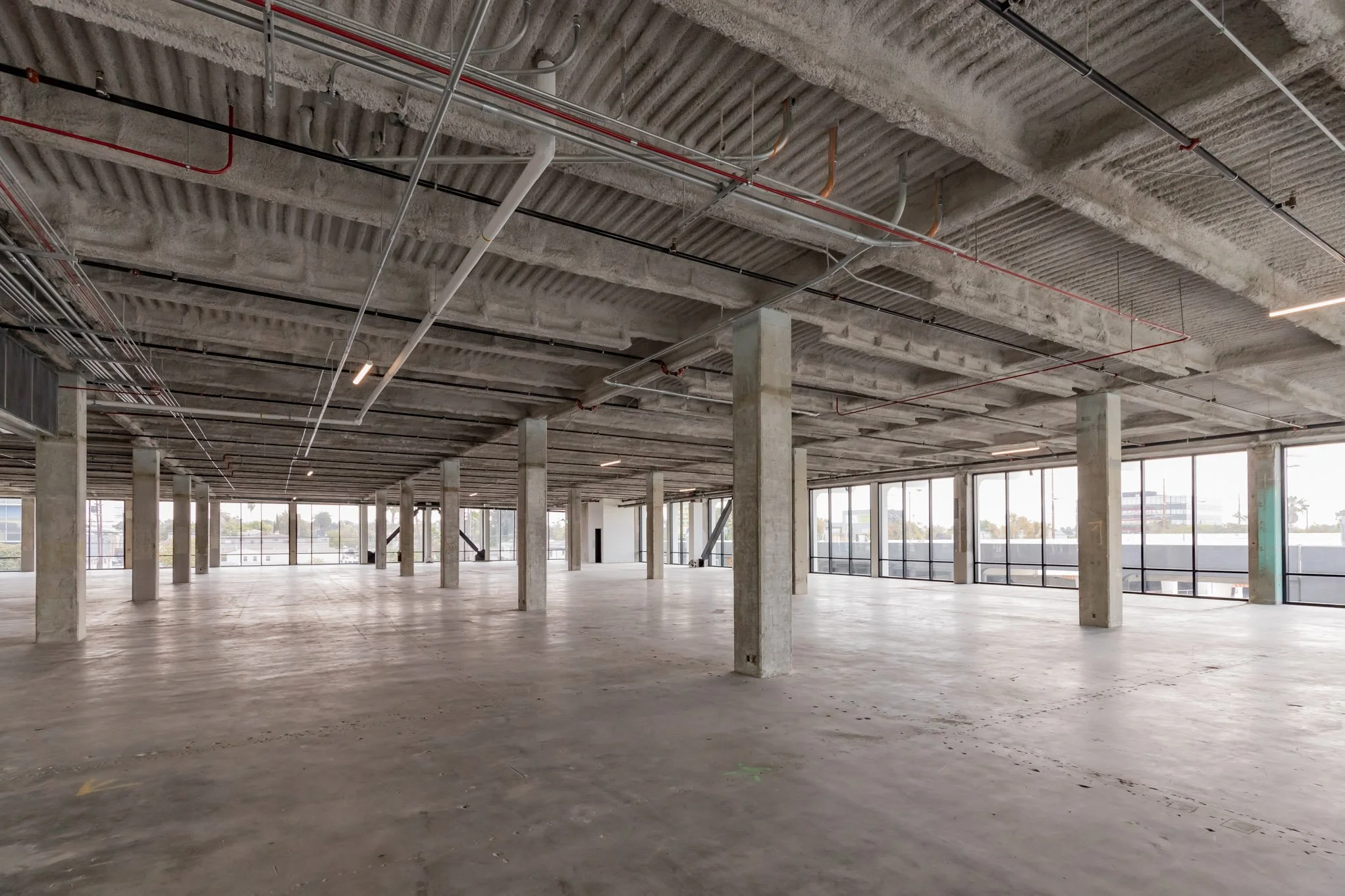 Empty commercial space with concrete floors, exposed ceiling pipes, and large windows allowing natural light