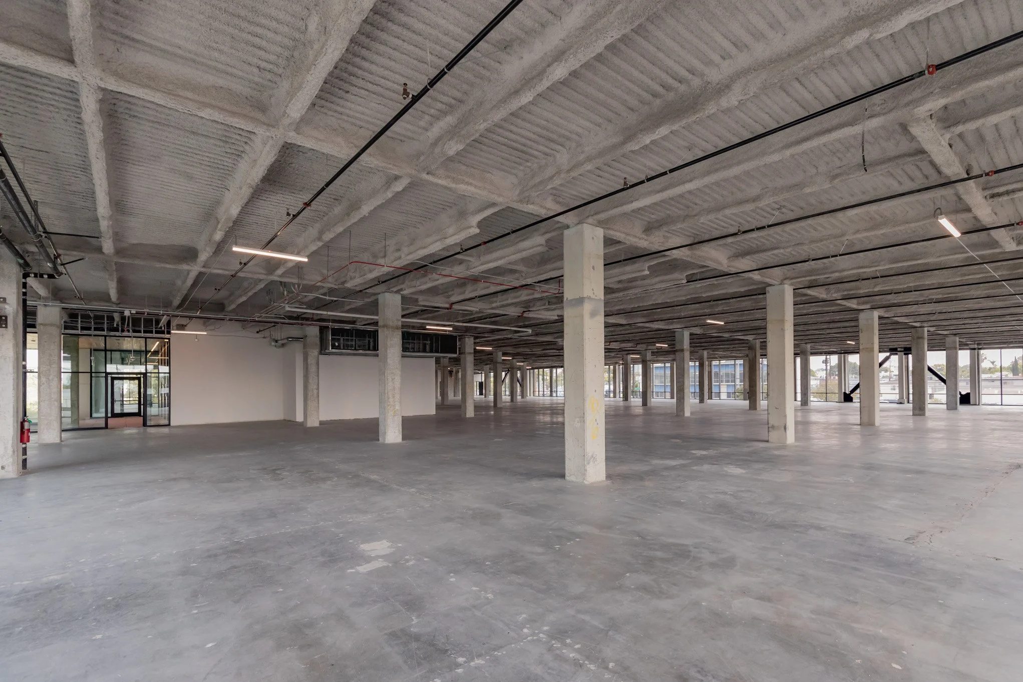 Empty unfinished commercial building with concrete floors, concrete support columns, exposed ceiling with electrical conduit, and large windows.