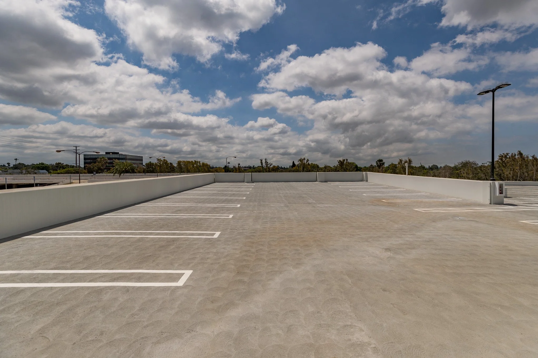 Empty rooftop parking lot with marked parking spaces, white wall barriers, tall street lights, and a partly cloudy sky overhead.