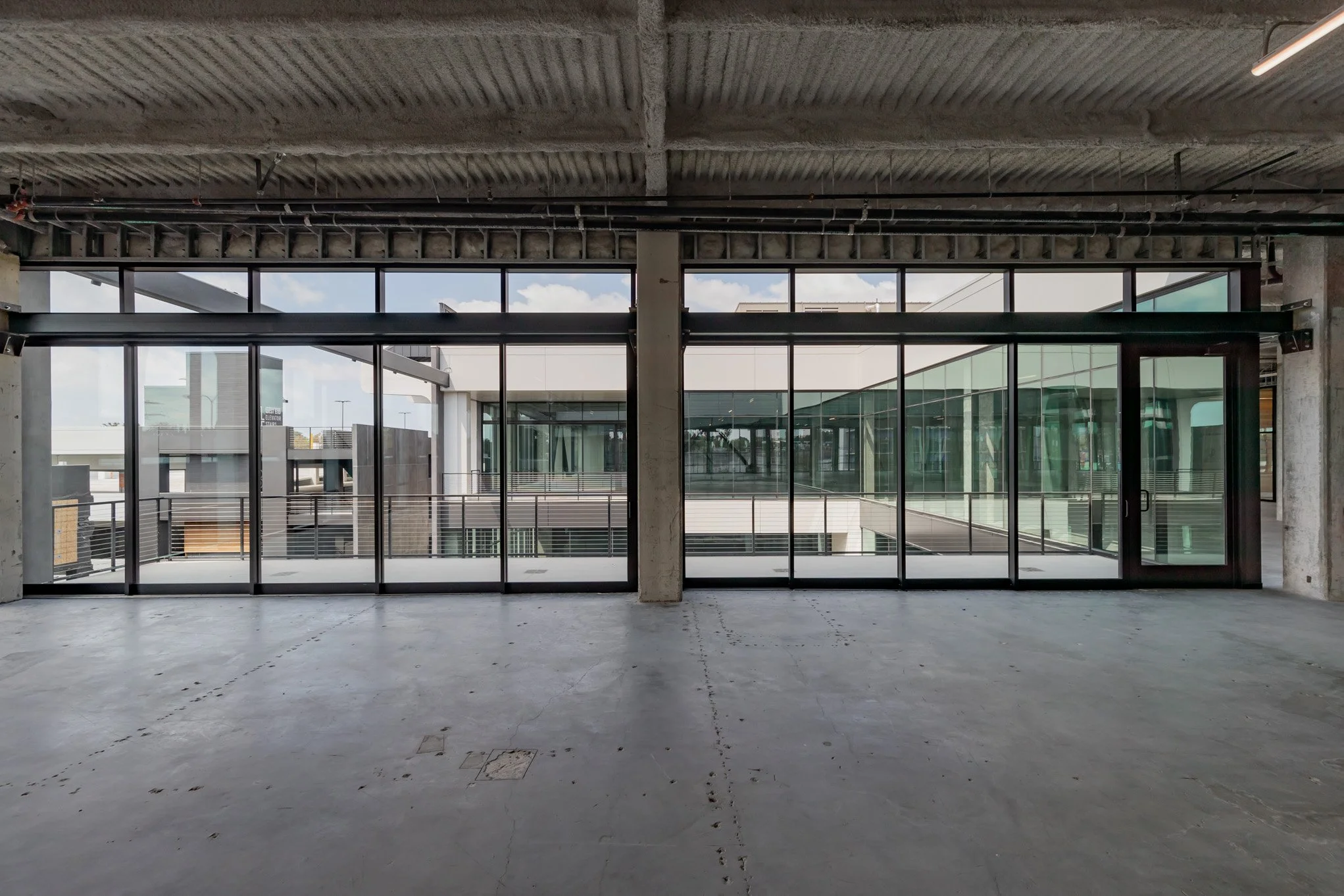 Empty unfinished interior space with large glass windows and doors, exposed ceiling, and view of the modern building's exterior.