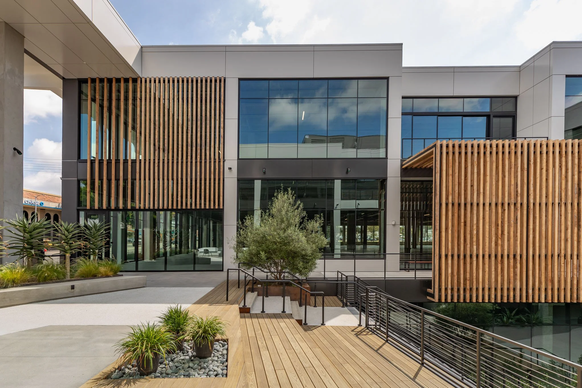 Modern building with gray and black exterior, large glass windows, and wooden slat accents; landscaped courtyard with plants, small trees, and a wooden platform with metal railings.