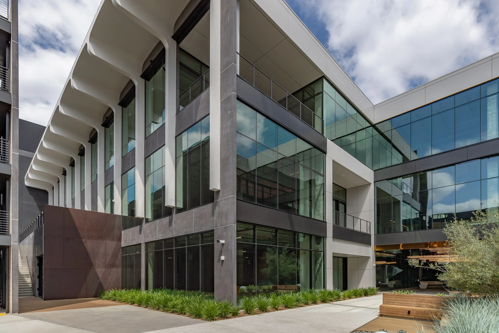 Modern multi-story office building with glass windows, concrete accents, and architectural white overhangs, surrounded by greenery and a pathway.