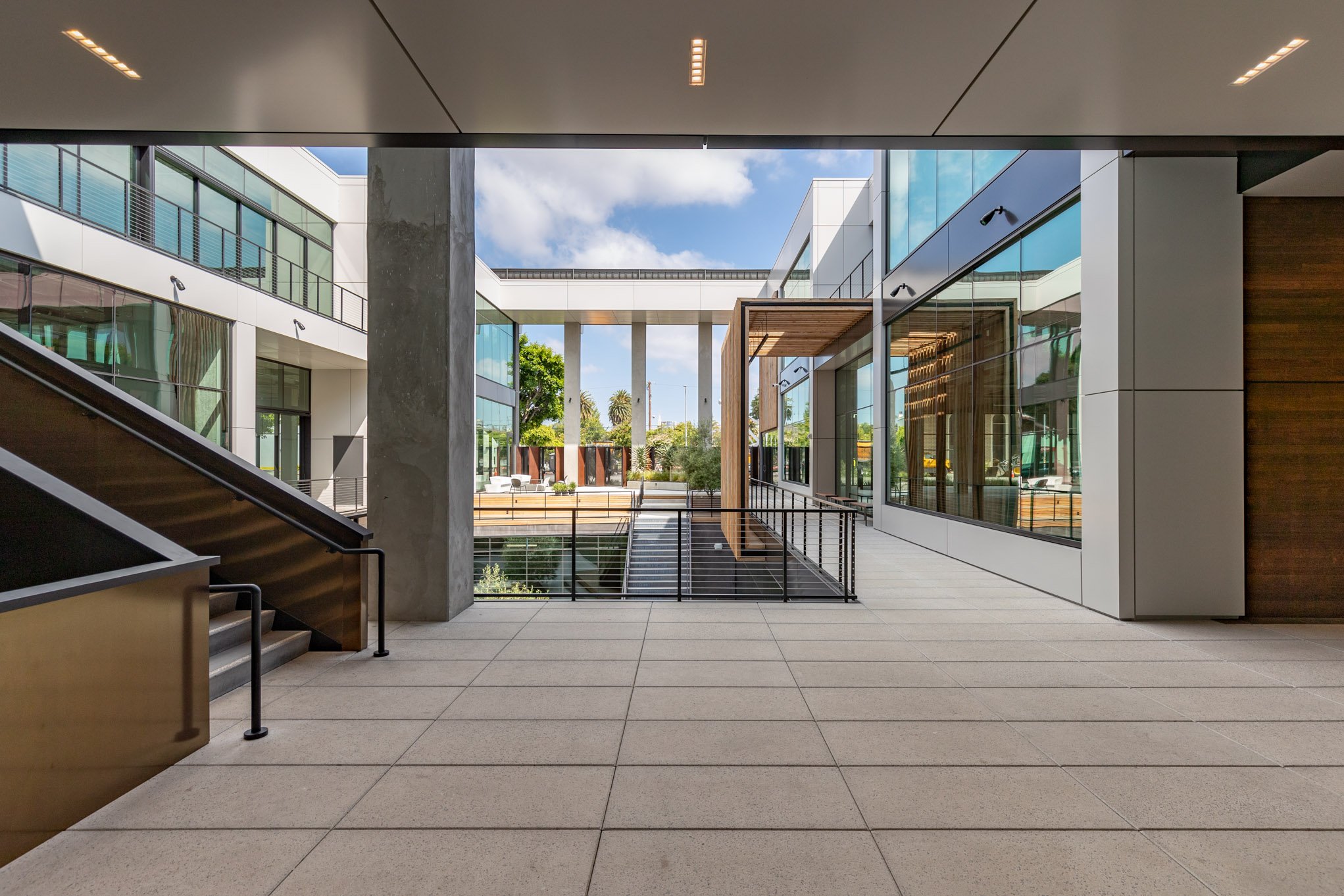 Modern commercial building interior with glass walls, tiled flooring, staircase, and outdoor patio area visible through large windows
