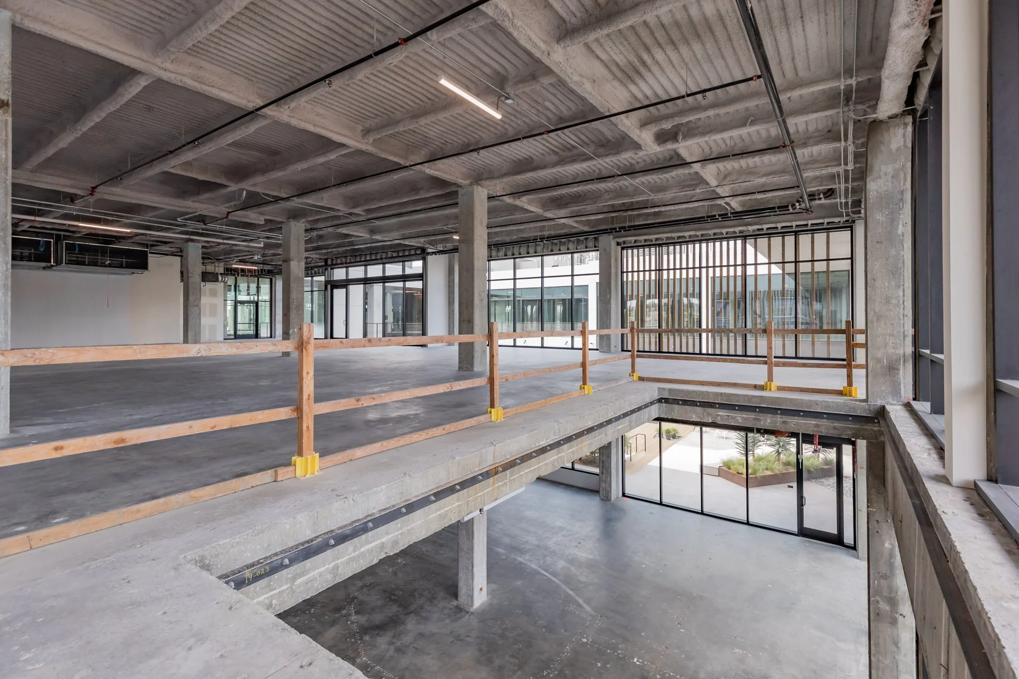 Interior view of an unfinished multi-story commercial building with exposed concrete, large windows, and construction barriers along the edges of the upper level.
