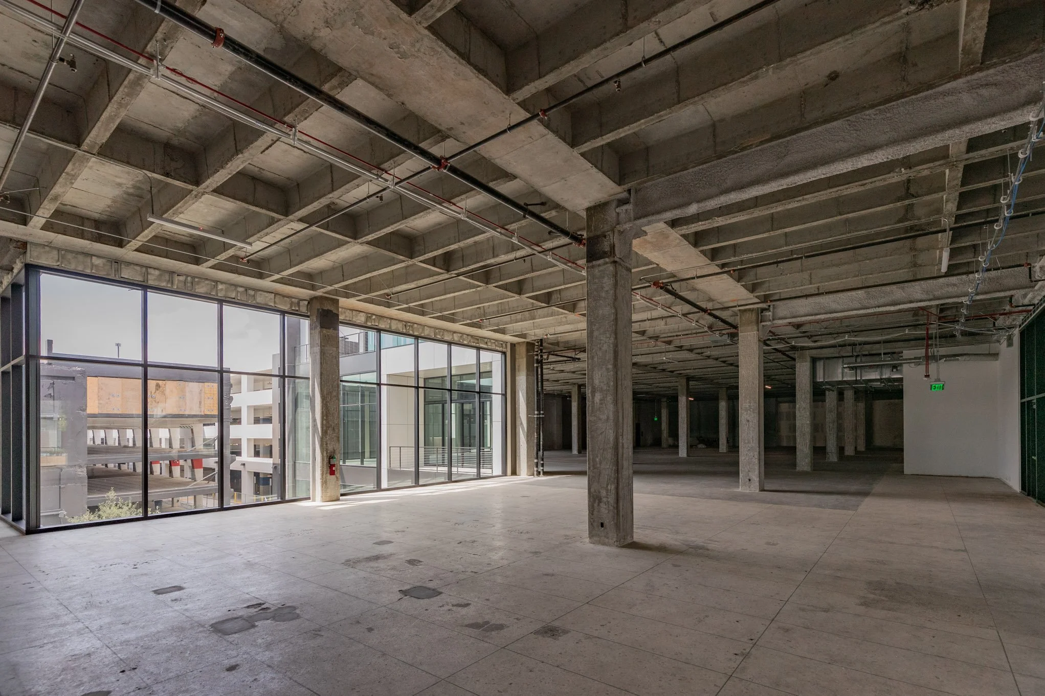 An empty, unfinished commercial building interior with concrete columns, exposed ceiling pipes, large glass windows, and tiled flooring.