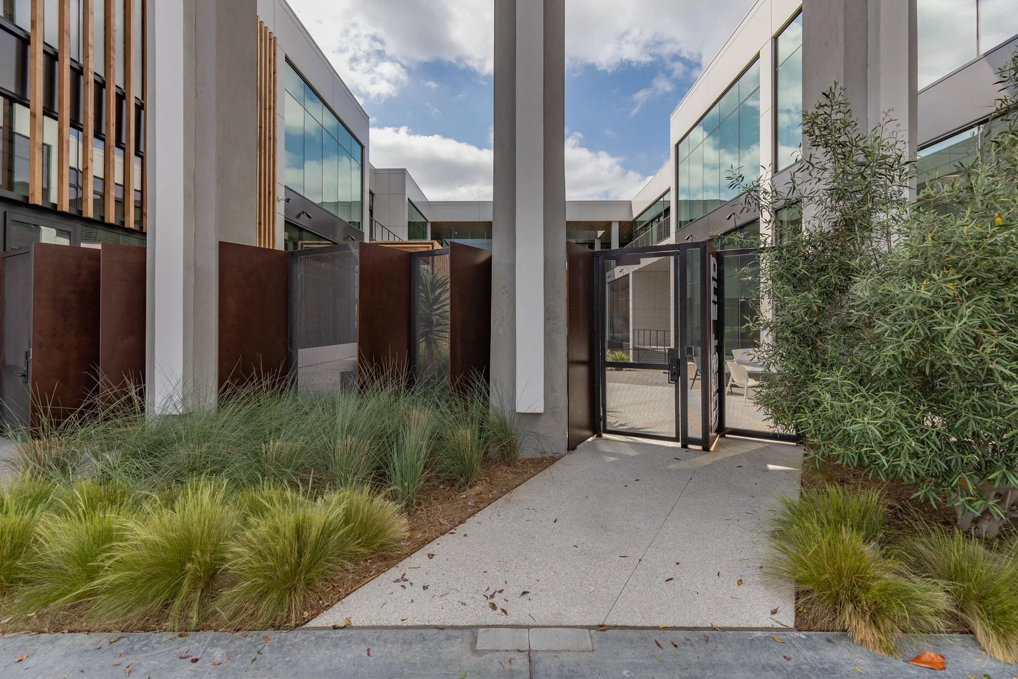 Modern apartment complex entrance with glass and metal fencing, concrete pathway, green plants, and a clear sky.