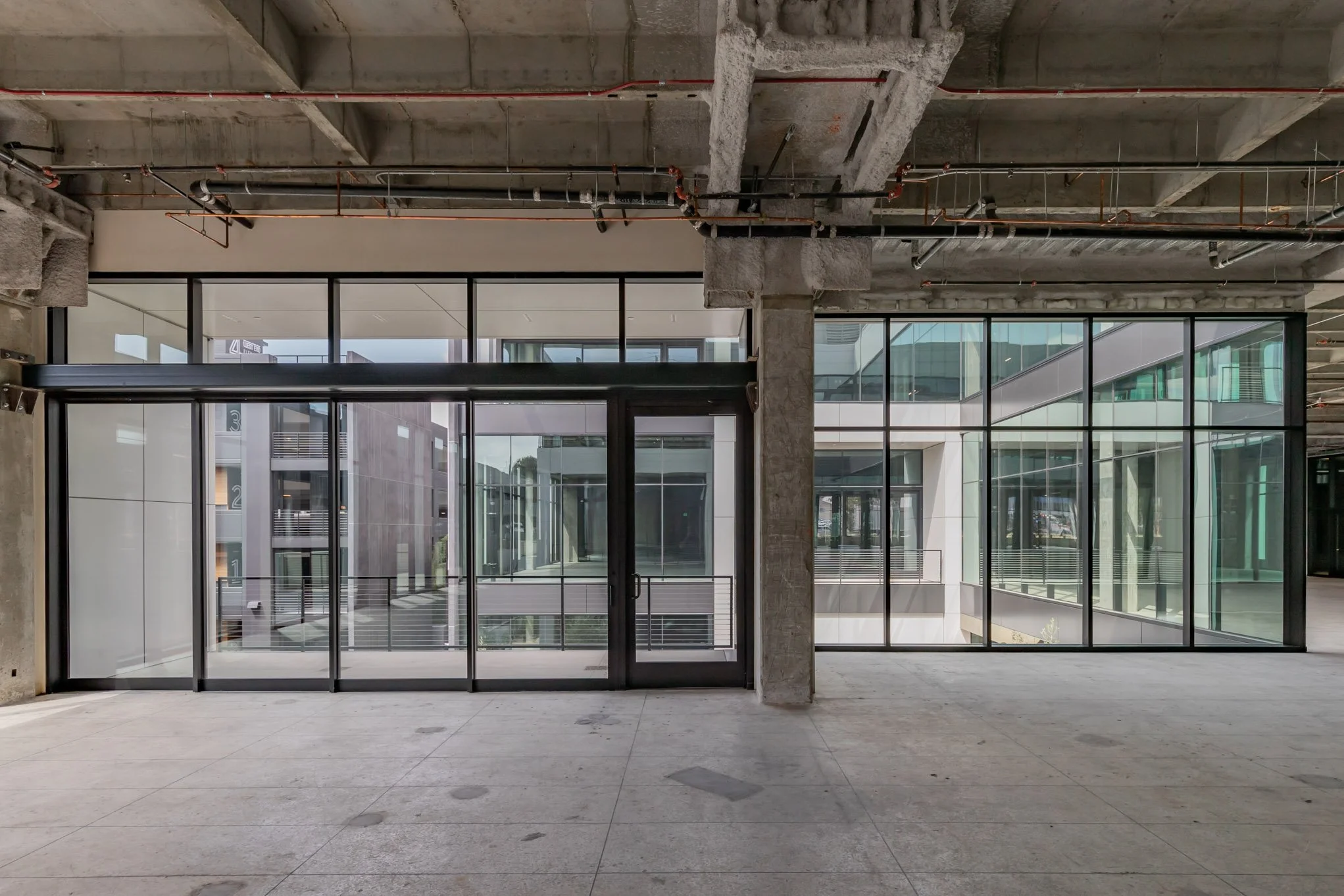 Interior of a modern, under-construction building with unfinished concrete ceiling, exposed pipes, and large glass windows, showing an inner courtyard or open space with surrounding buildings.