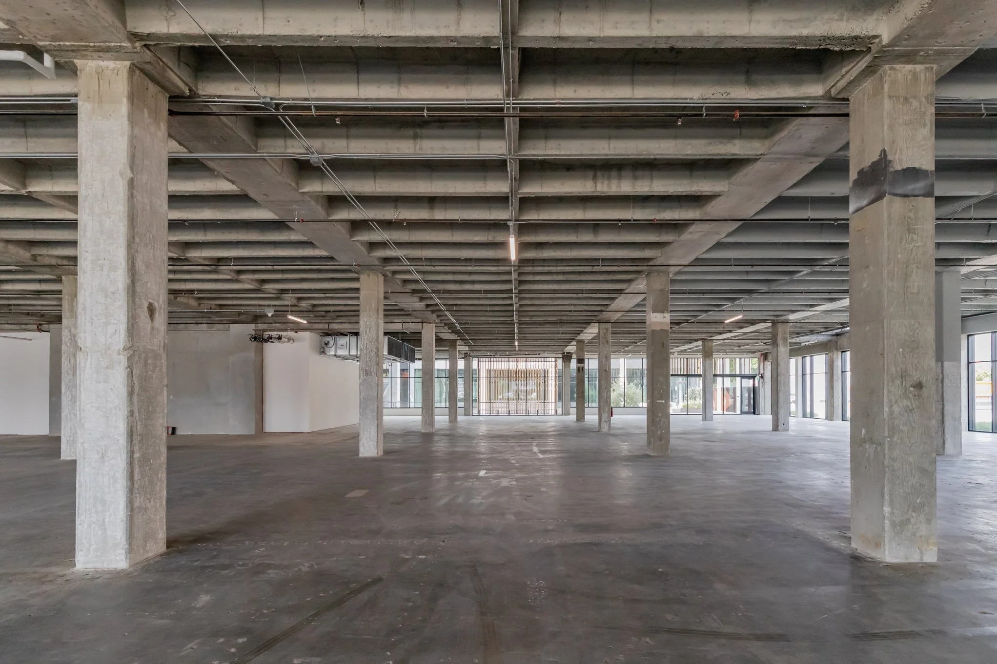 Empty, unfinished commercial space with concrete columns, exposed ceiling, and large windows.