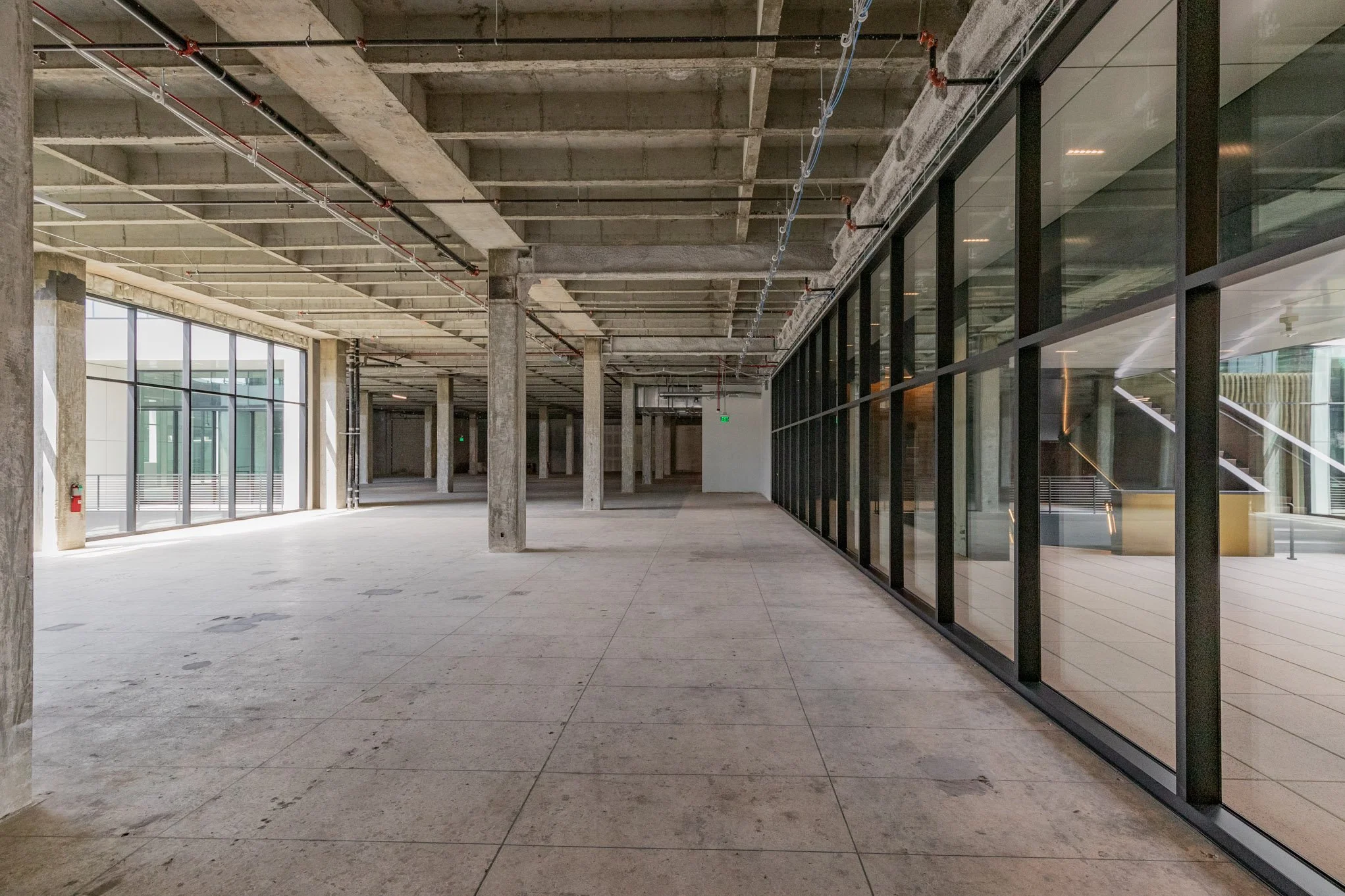 Empty interior space of a modern building under construction with concrete floors and ceilings, exposed pipes, and large glass windows.