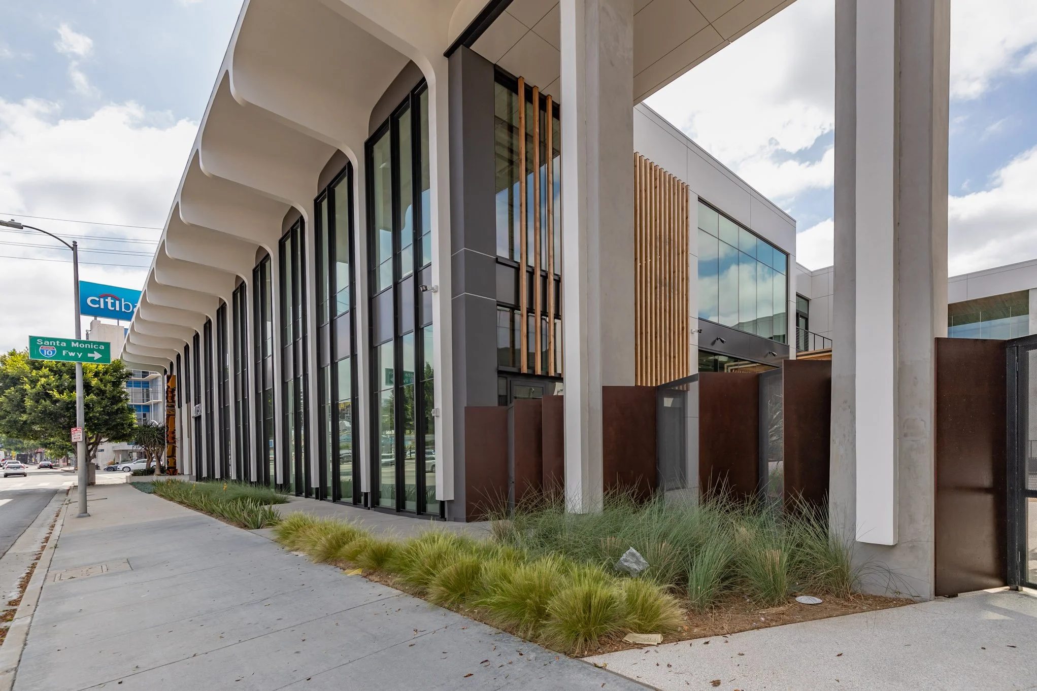 Modern building with large glass windows, concrete and wood accents, on a city sidewalk with street signs for Santa Monica Freeway and CitiBank visible in the background.