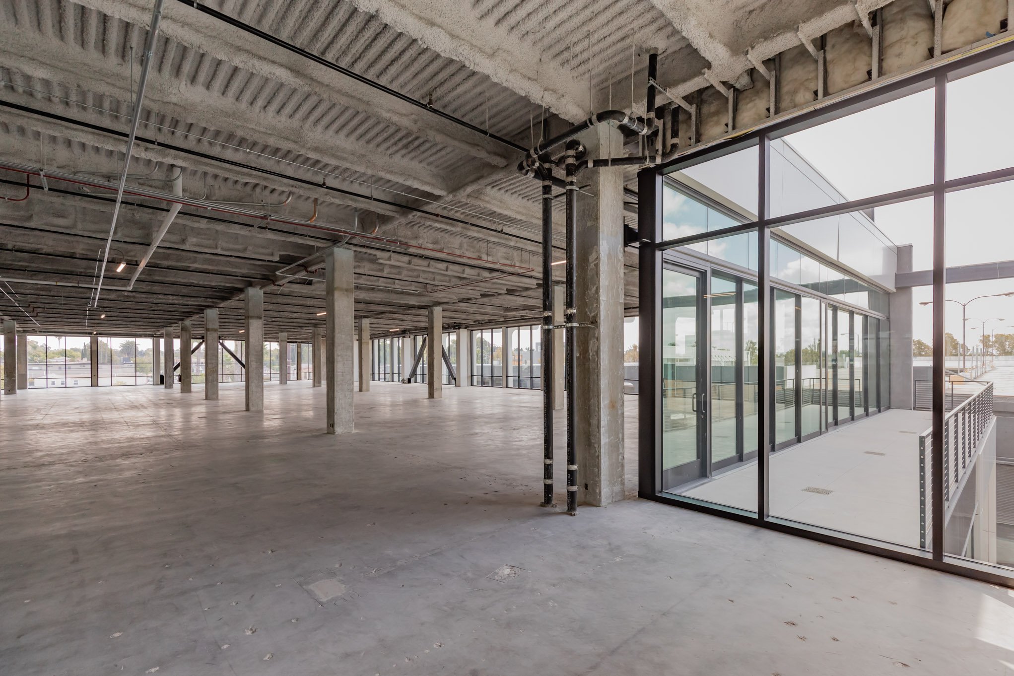 Empty, unfinished commercial space with exposed concrete floors, ceiling, and support columns, large glass windows and doors, and outdoor balcony access.