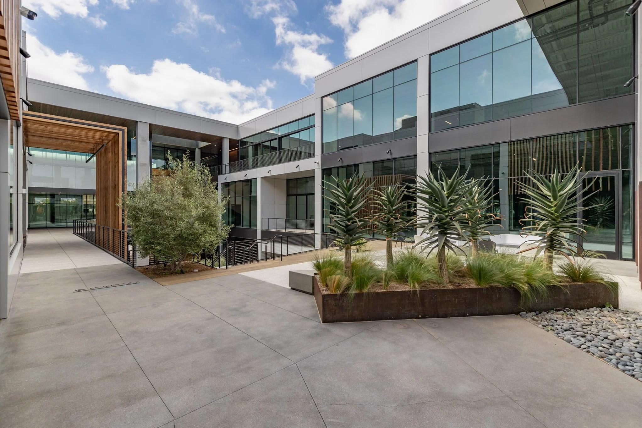 Modern office building courtyard with large glass windows, potted plants, a small tree, and a concrete walkway under a partly cloudy sky.
