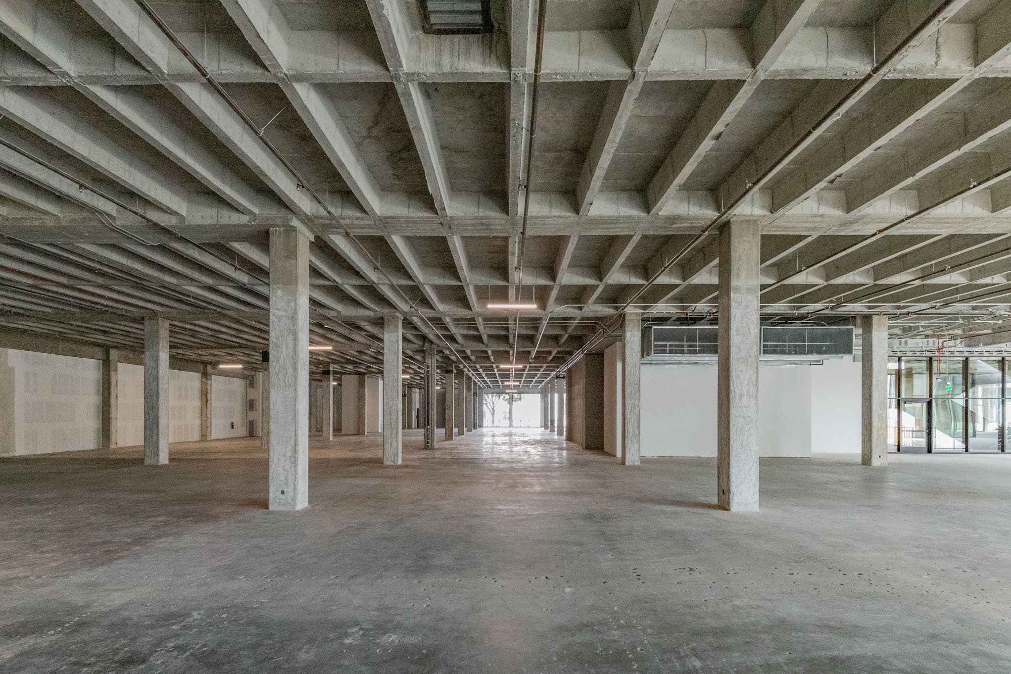 Empty concrete commercial space with exposed ceiling beams, support columns, and large front windows