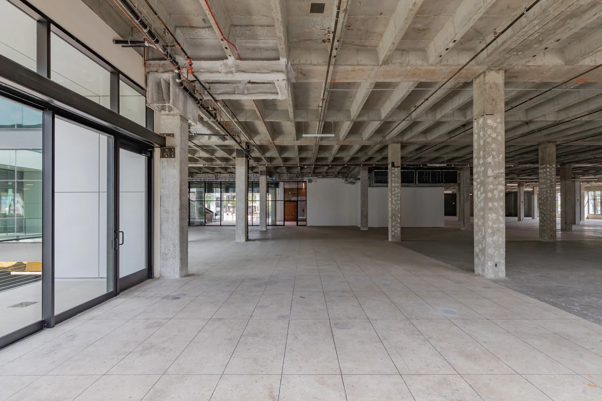 Empty commercial or office space with concrete columns and ceiling, tiled floor, glass walls and doors, and exposed ceiling pipes.