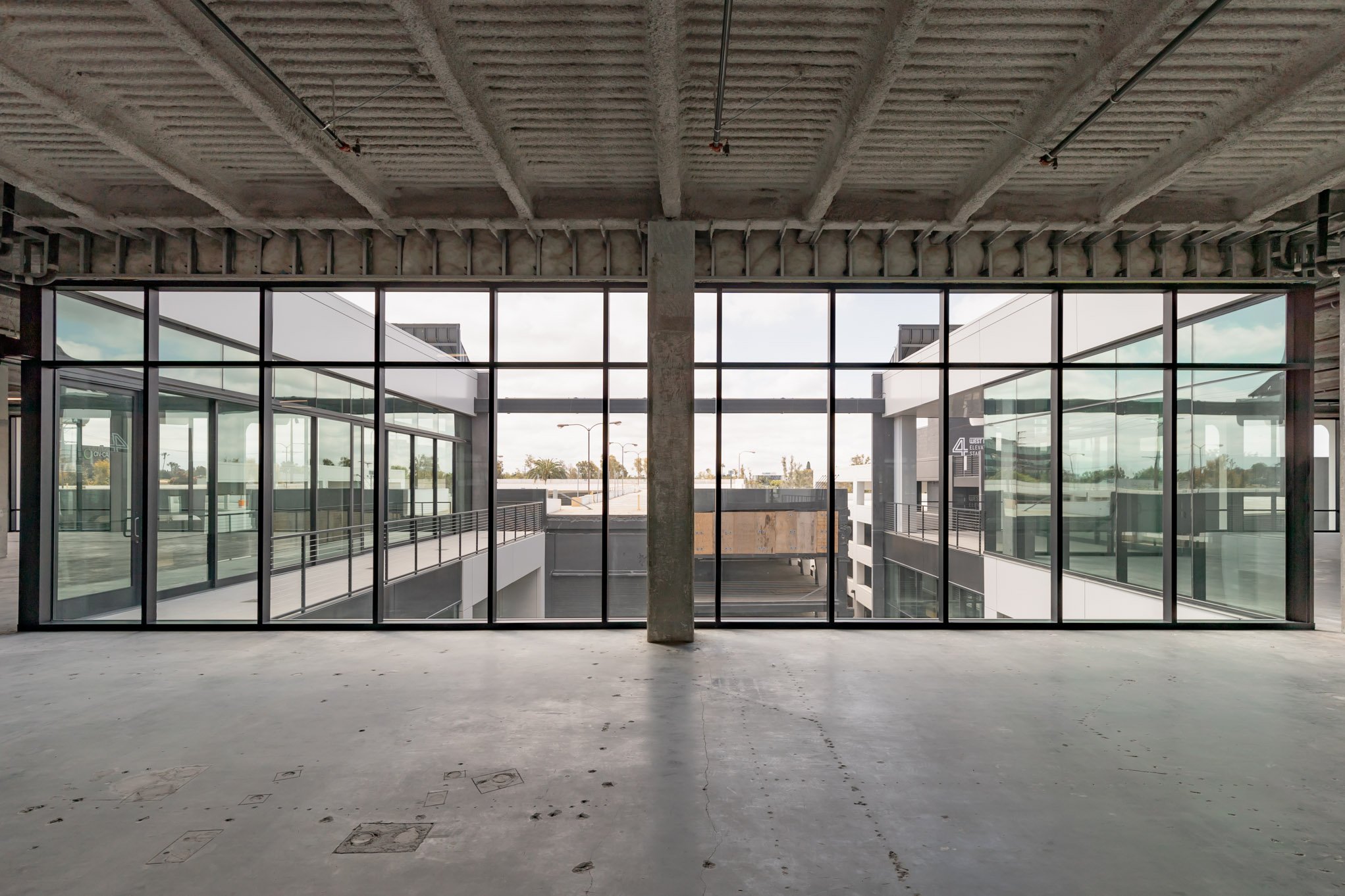 Empty unfinished building interior with large glass windows overlooking a parking lot and cityscape.