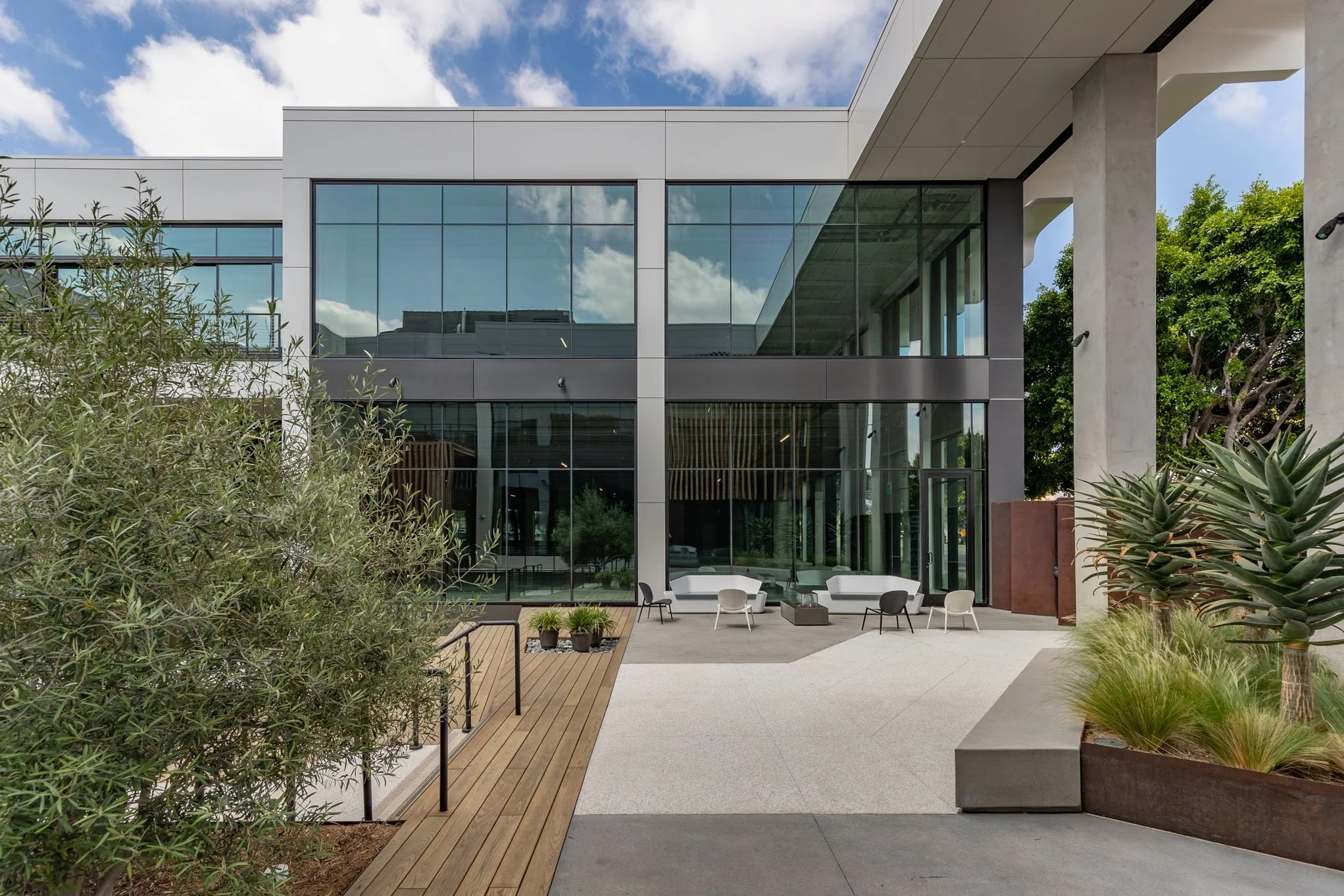 Modern office building with large glass windows, outdoor seating area with chairs and sofas, surrounded by greenery and trees, under a partly cloudy sky.