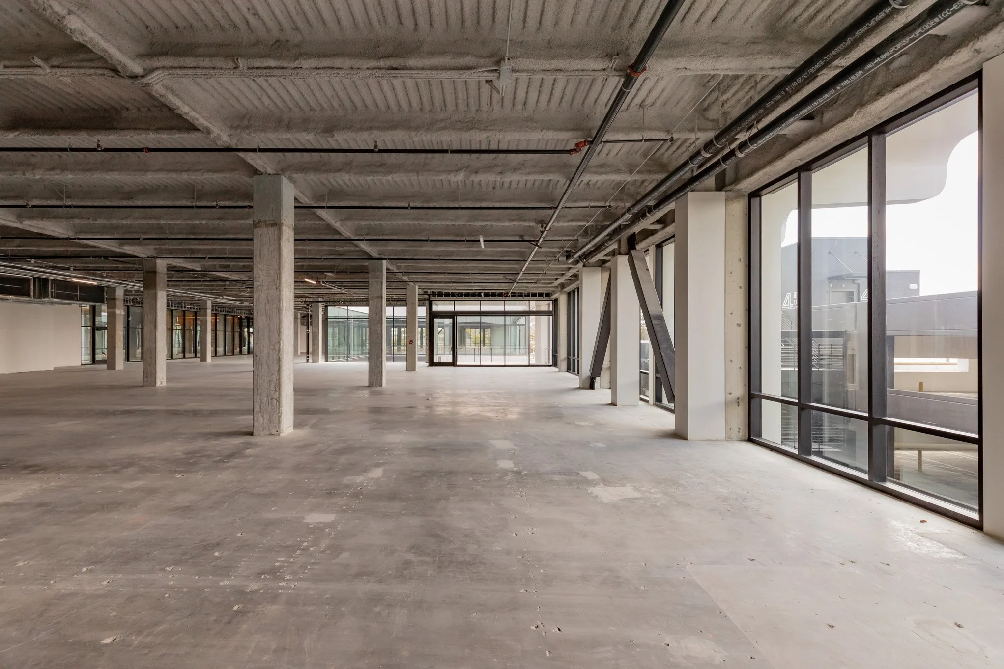 Empty commercial building space under construction with concrete floors, support columns, exposed ceiling with pipes, and large floor-to-ceiling windows.