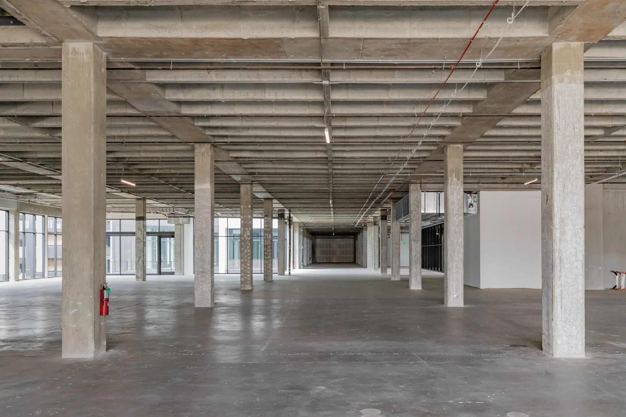 Empty, unfinished commercial interior space with concrete pillars, exposed ceiling beams, and large windows.