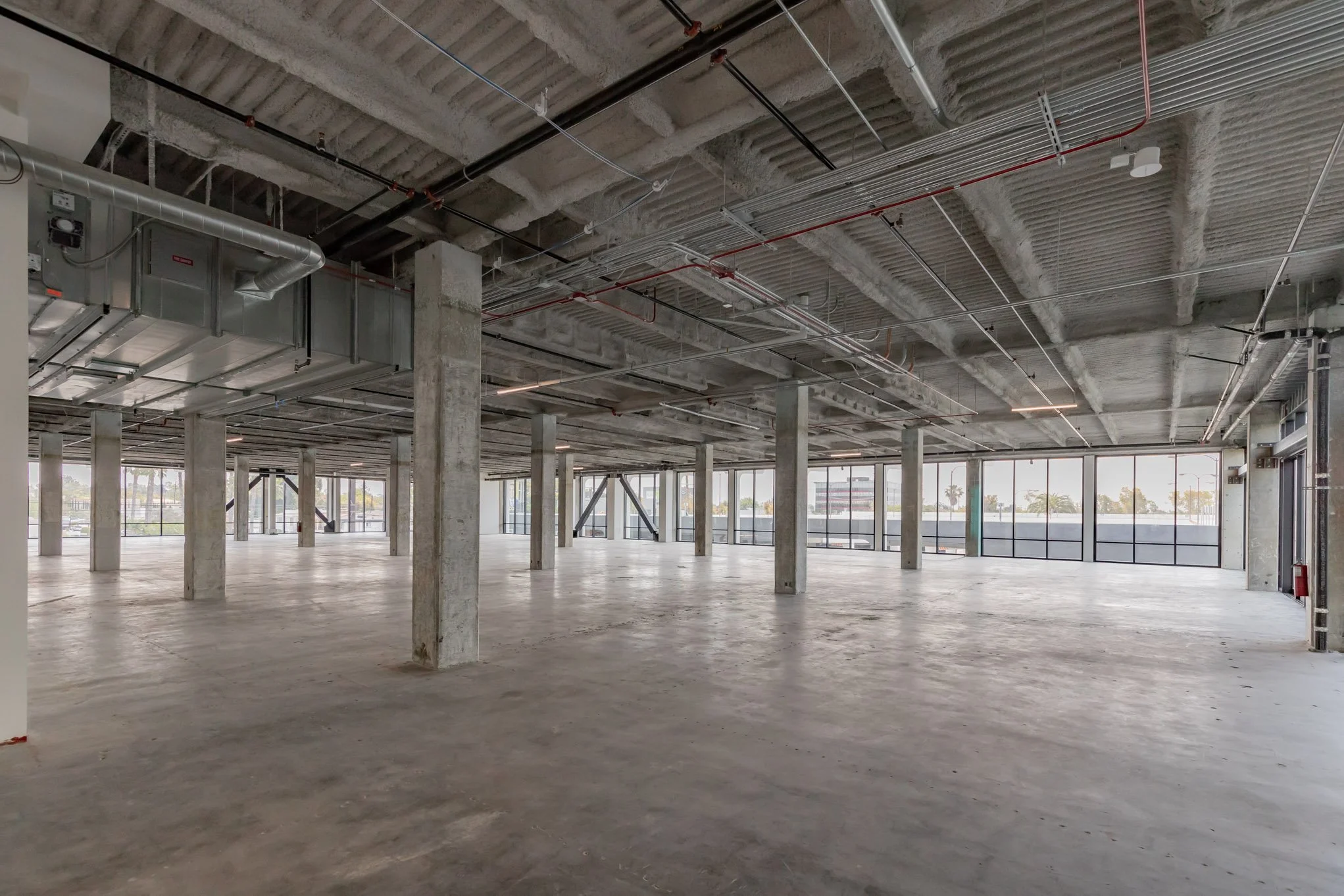 Empty commercial space under construction with concrete pillars, ductwork, and large windows.