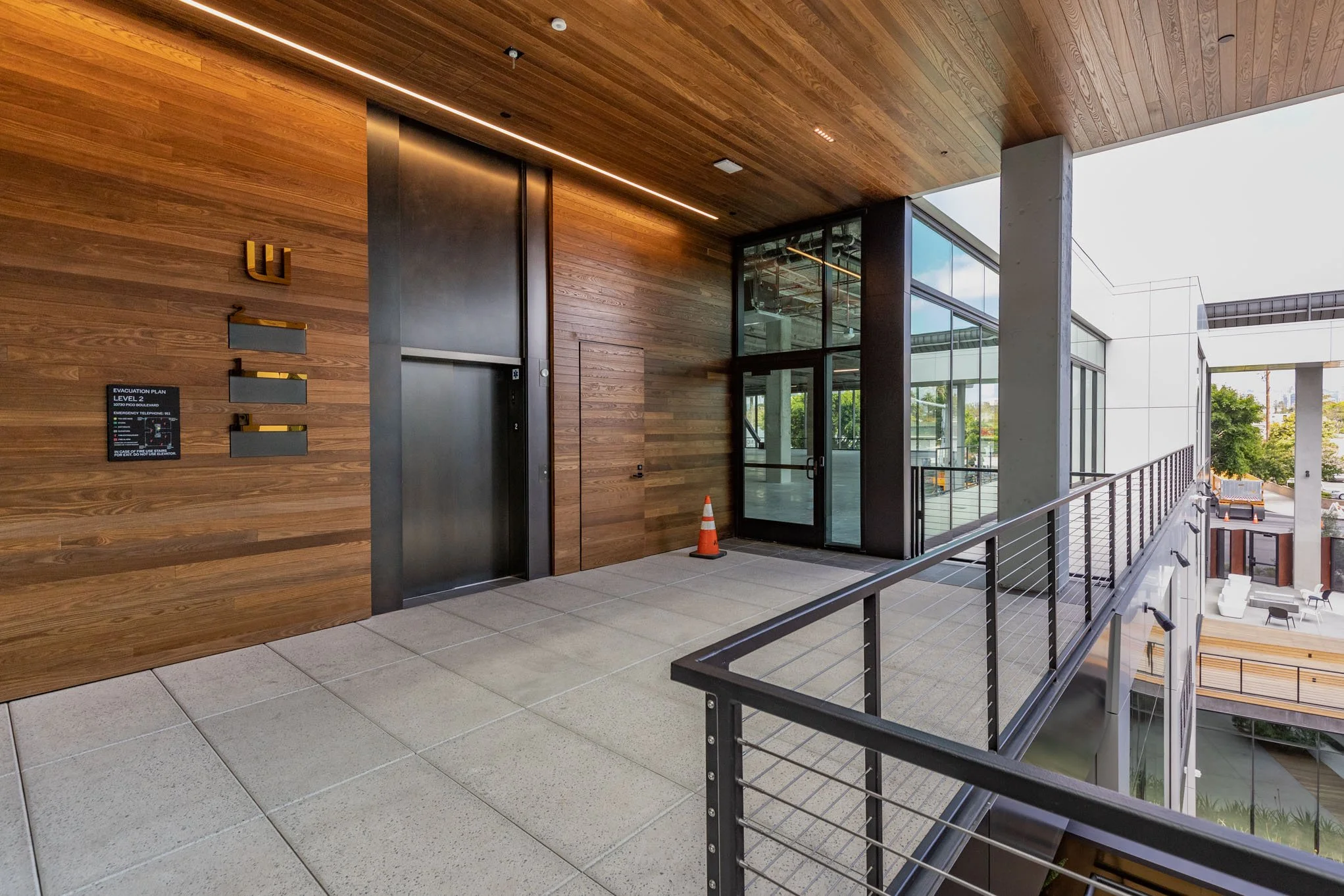 Modern multi-level building hallway with wooden walls, glass doors, and an elevator, featuring a safety cone and outdoor patio area with seating.