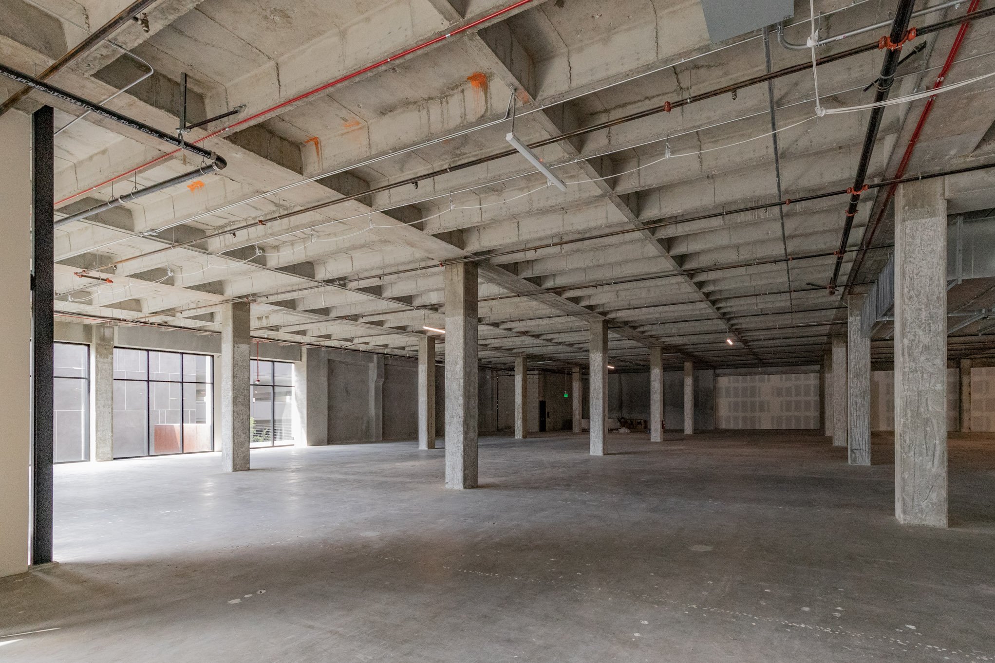 Empty commercial building interior with concrete floors, square concrete support columns, and unfinished ceiling with exposed pipes and ductwork.