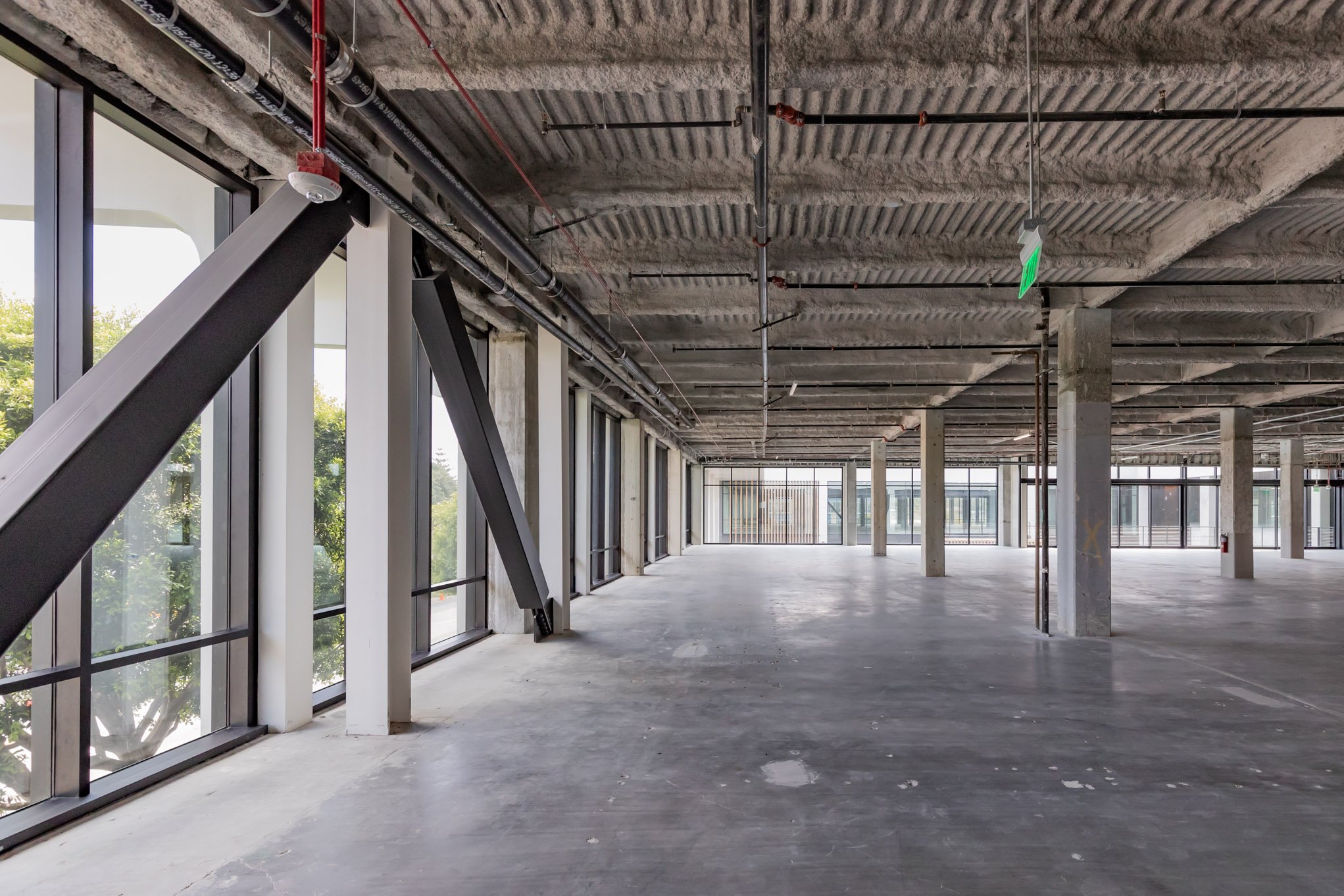 Unfinished interior space with concrete floors, exposed ceiling with insulation, metal pipes, and large windows with view of trees.