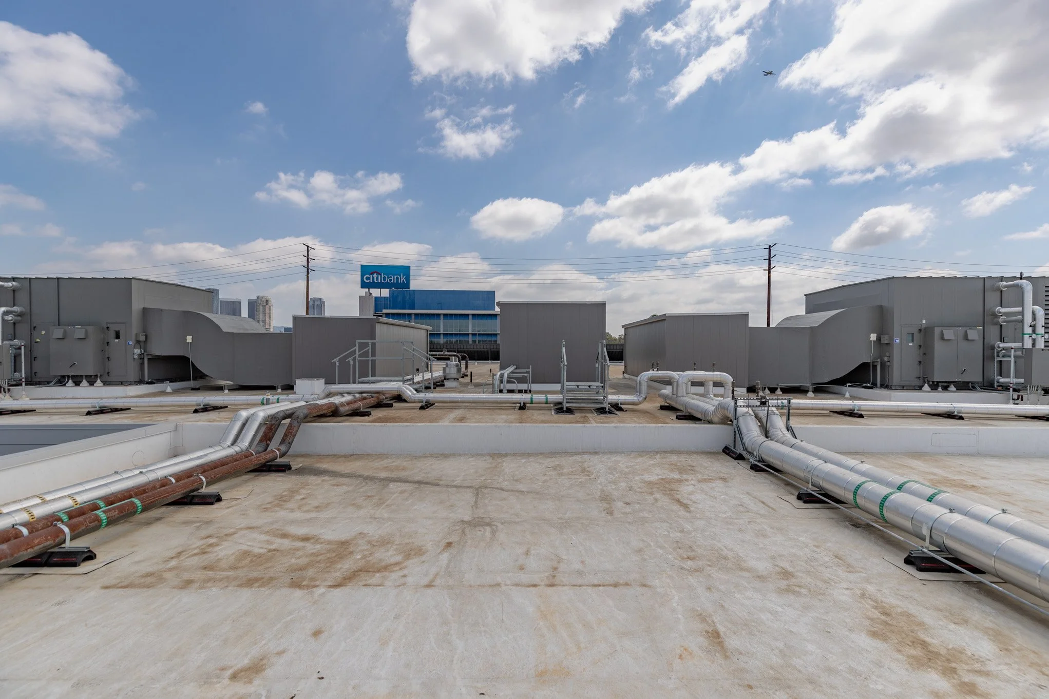 Rooftop with industrial HVAC units and ventilation ducts, blue sky with clouds, city skyline in the background, and an airplane flying overhead.
