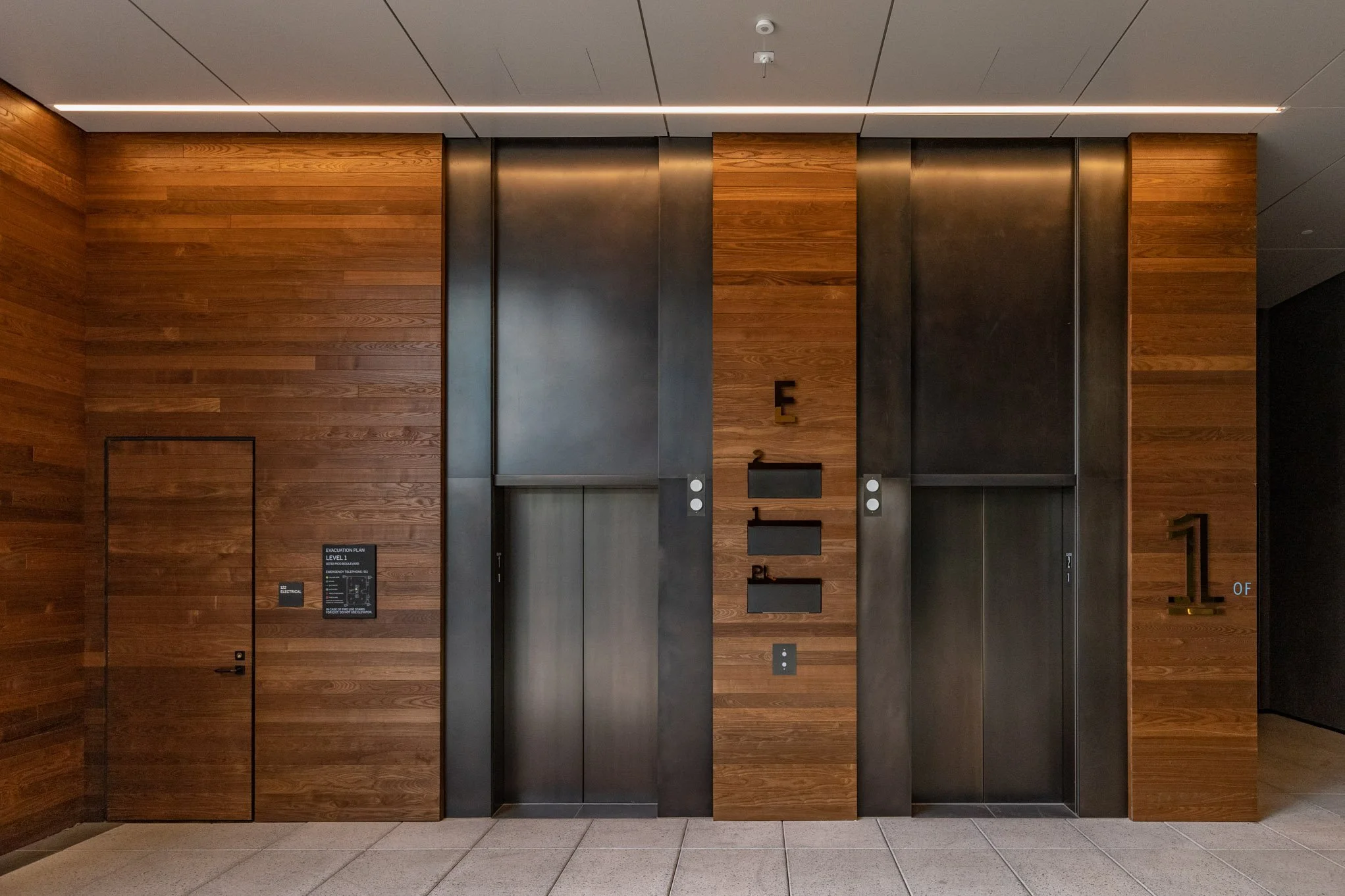 Interior view of a modern building with two black elevator doors framed by wood paneling, with a fire evacuation plan on the wall to the left of the elevators and a large gold number 1 indicating the floor.