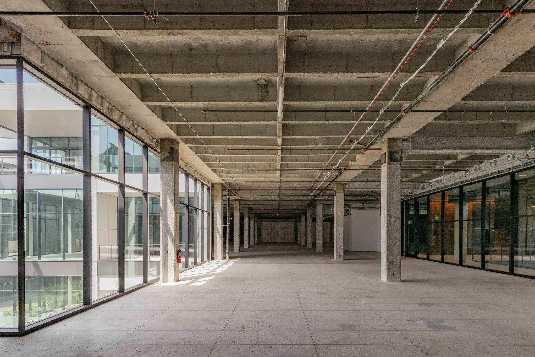 Empty interior space of a building under construction with large glass windows, concrete columns, and exposed concrete ceiling.
