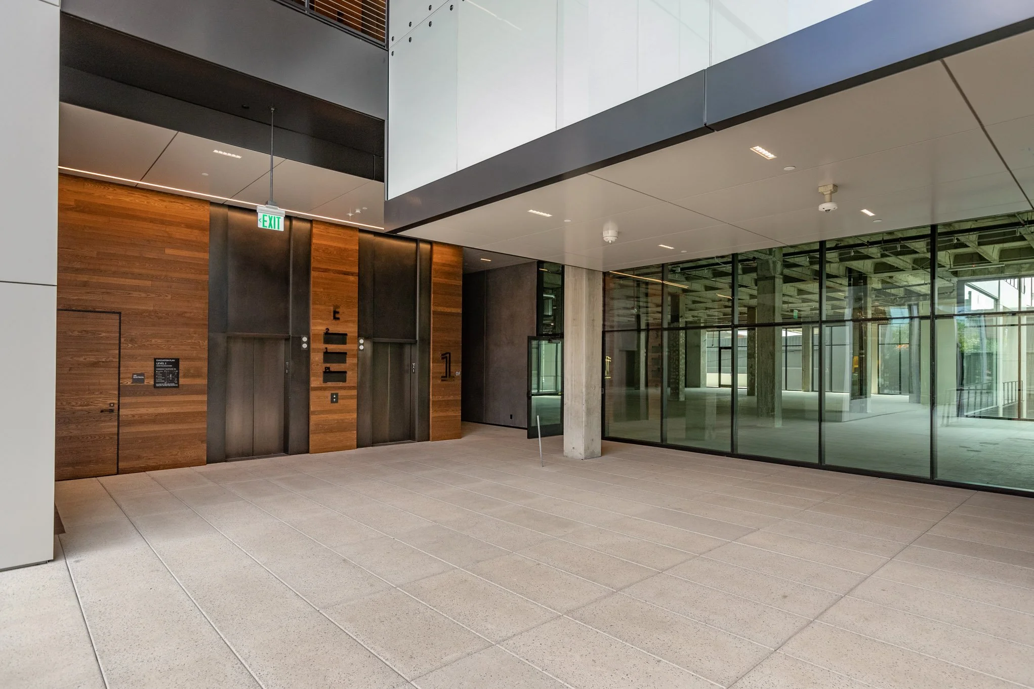 Interior of a modern building lobby with two elevators, wood paneling, glass walls, an exit sign, and minimal decor.