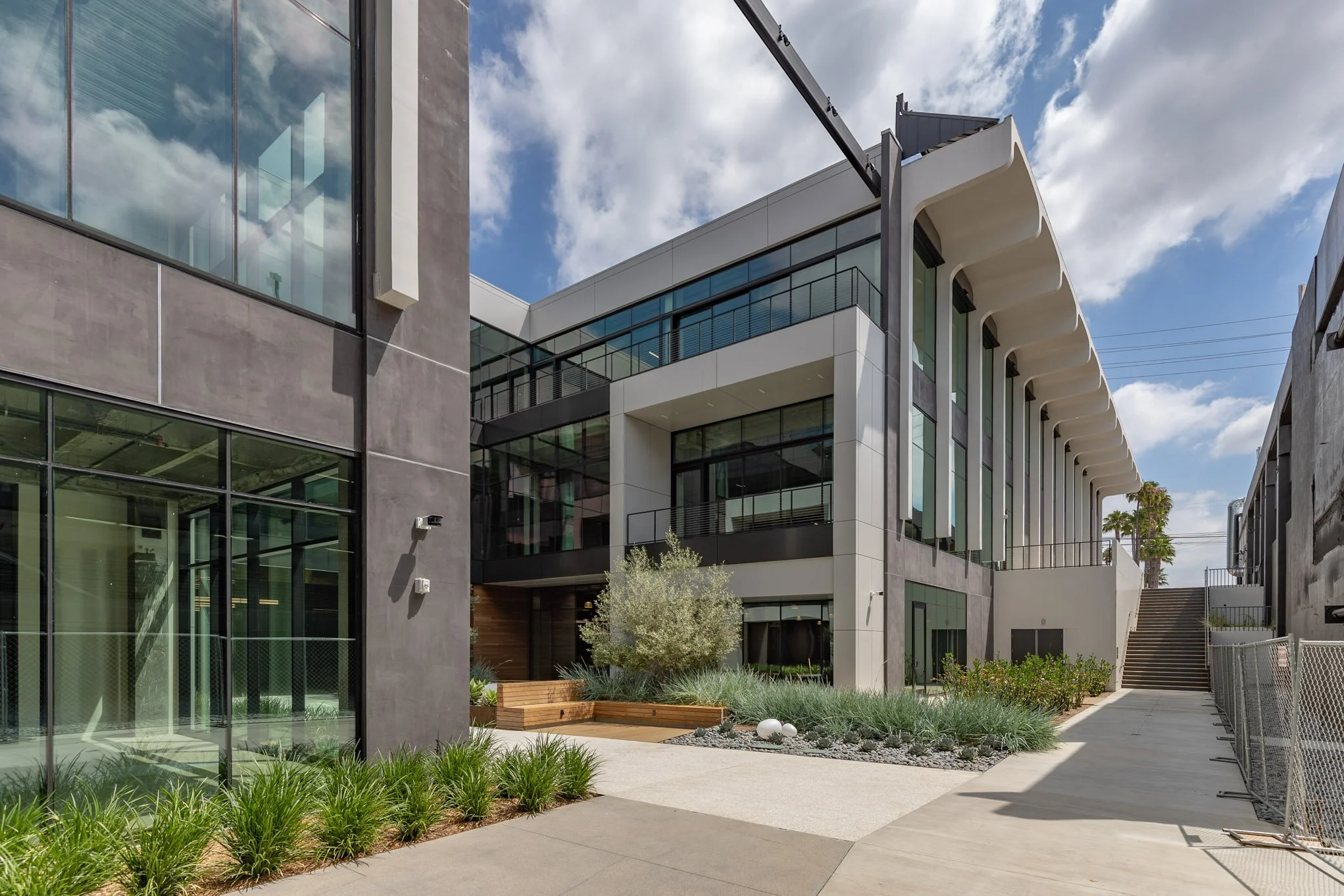 Modern multi-story building with glass windows and a landscaped outdoor area, showing stairs, plants, and a blue sky with clouds.