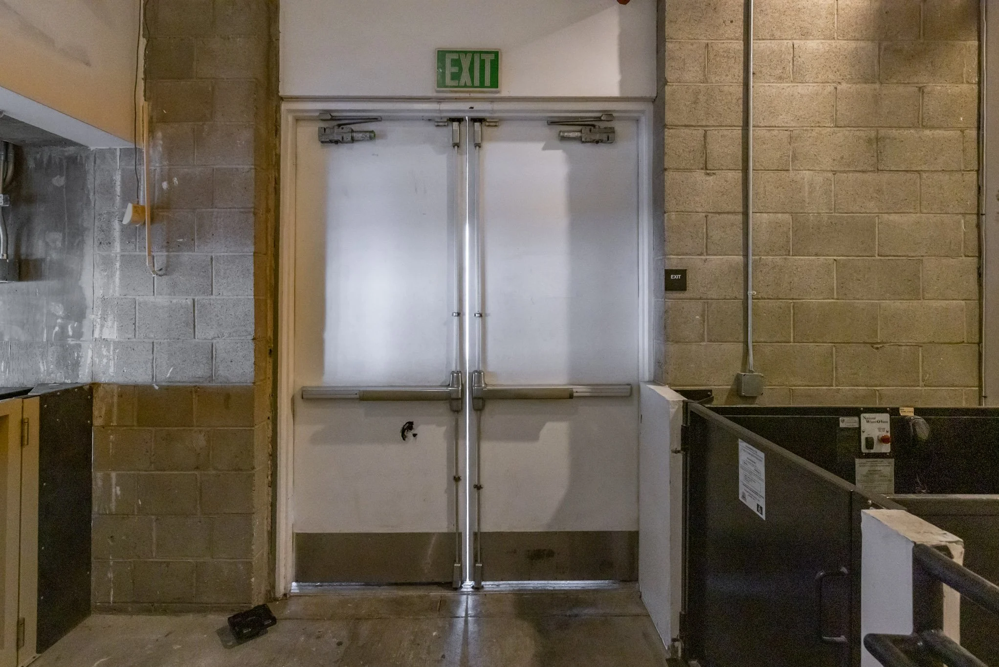 Double metal emergency exit door with green 'EXIT' sign above, surrounded by brick and concrete walls, with some machinery and debris nearby.