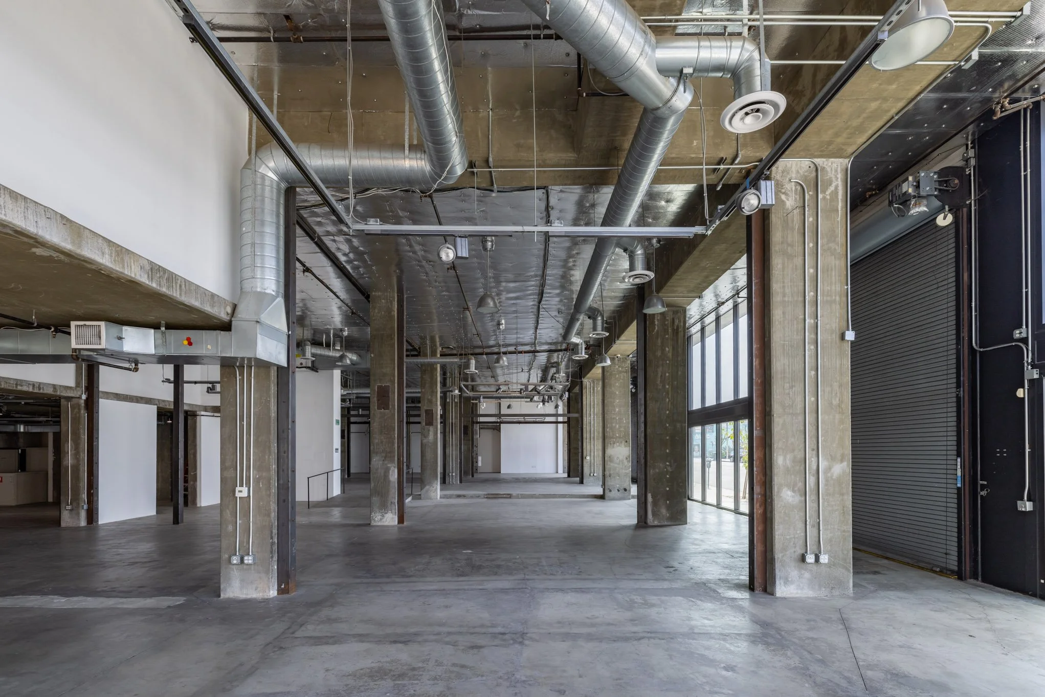 Empty commercial warehouse space with exposed concrete columns, ductwork, and a large roll-up door.