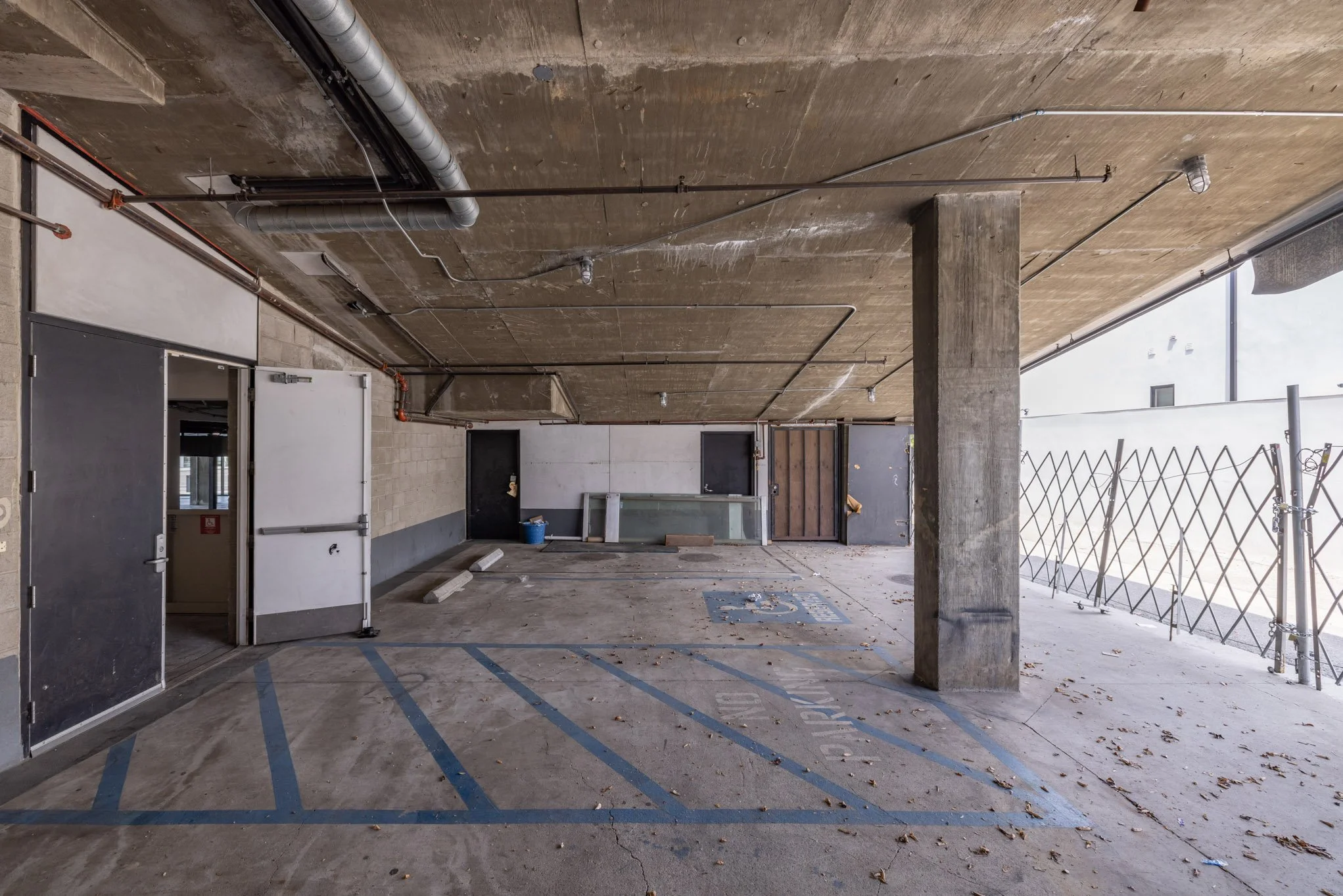 Empty parking space in a garage with a marked handicapped spot, concrete floor and ceiling, exposed pipes, and a concrete pillar, with debris on the ground and a partly enclosed balcony area to the side.