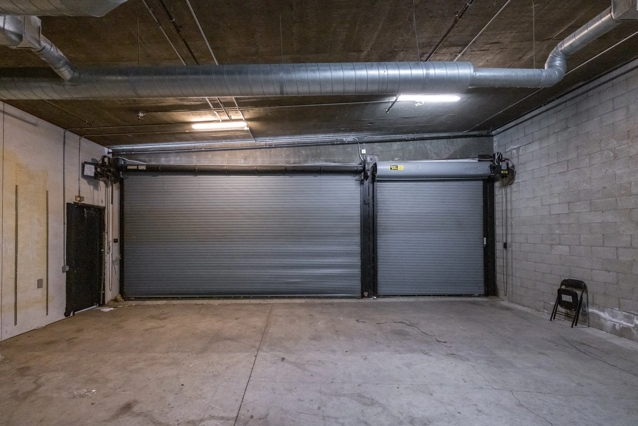 Empty indoor parking garage with two closed metal roll-up doors, concrete floor, concrete block wall, exposed pipes on ceiling, small black folding chair in corner.