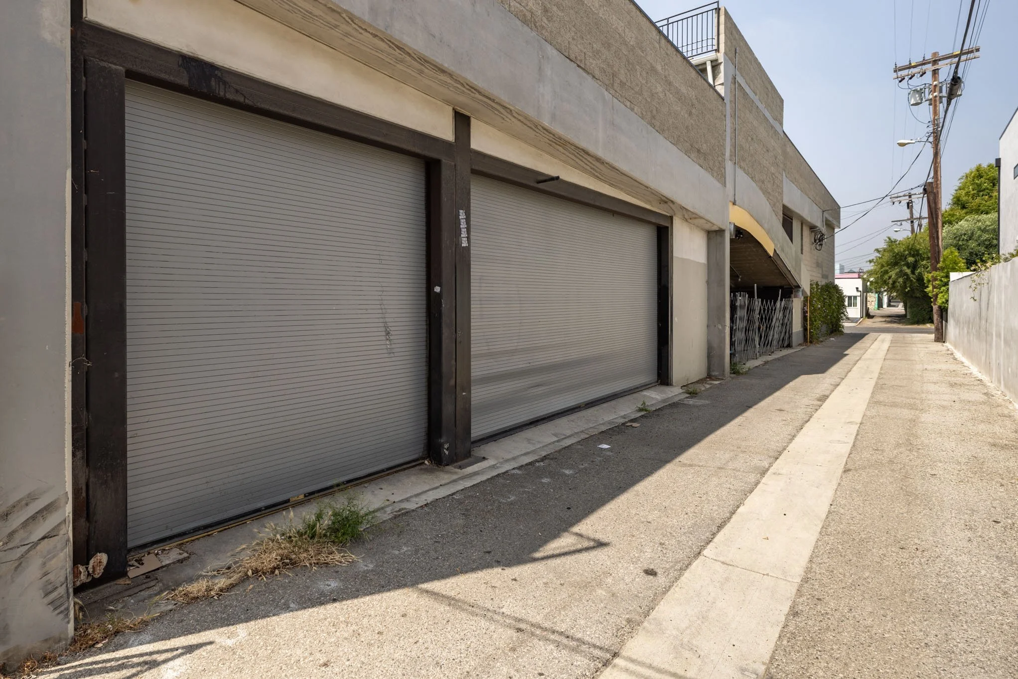 Empty sidewalk with a building with two closed roller shutters on the left side, utility poles and wires on the right, and trees and buildings in the background under a clear sky.