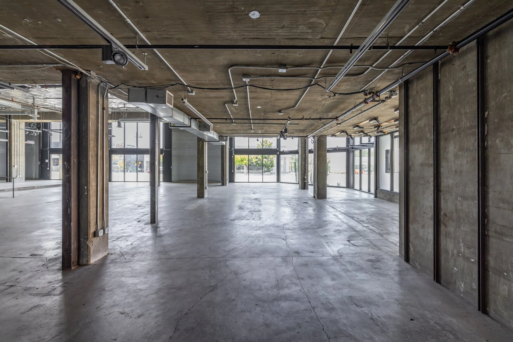 Empty commercial space with large windows, exposed concrete walls, ceiling with visible ducts and pipes, and polished concrete floor.