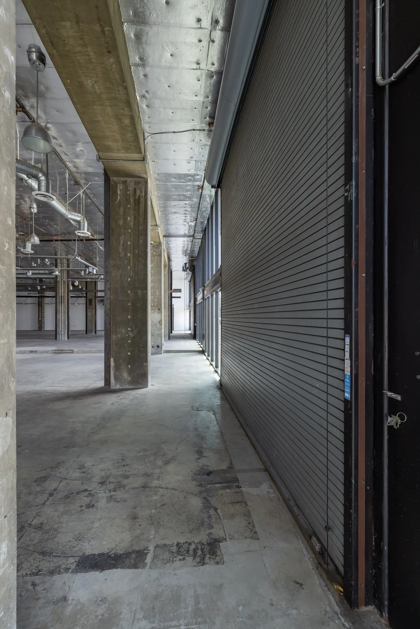 Inside an unfinished commercial building with concrete walls, ceiling, and floors, featuring exposed ductwork and a large metal roll-up door.