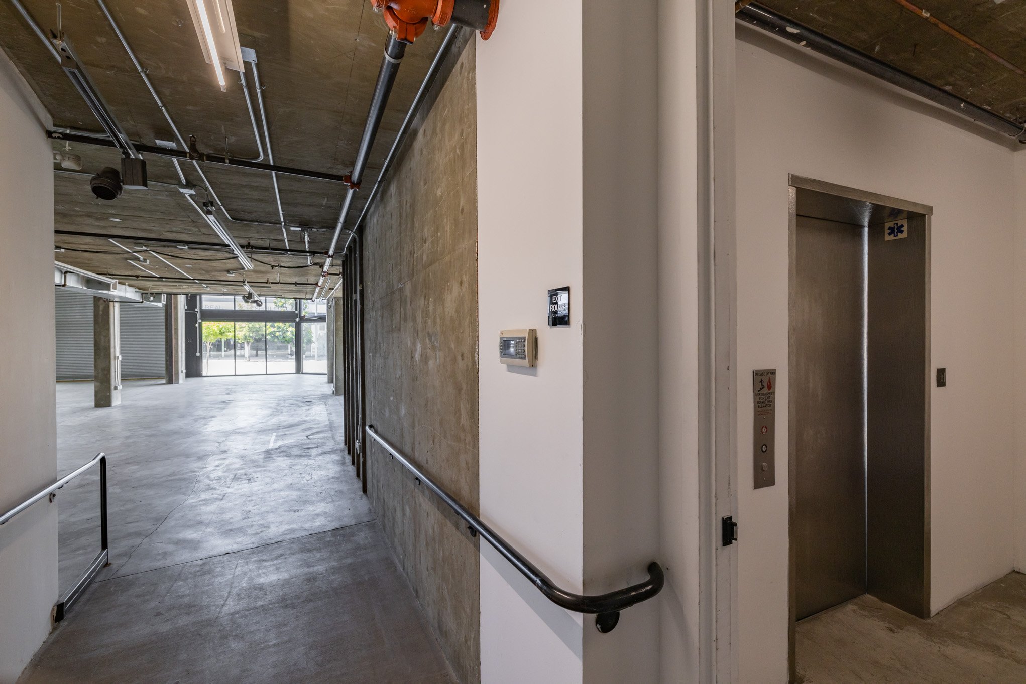 Empty commercial space with an elevator on the right, parking garage entrance at the end, and white walls and ceiling with visible piping.