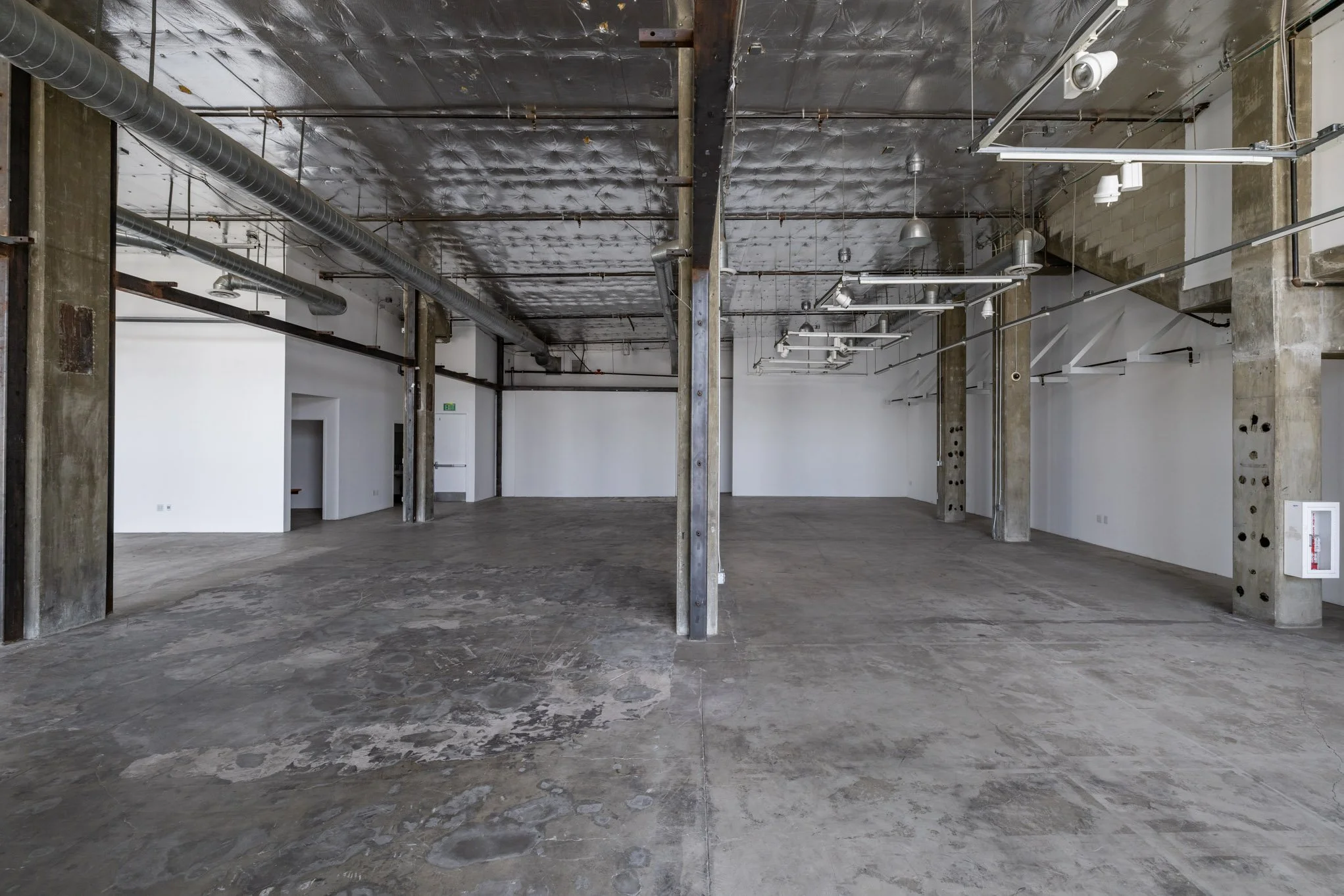 Empty industrial style space with concrete floors, exposed ducts, and metal beams, split into two sections.