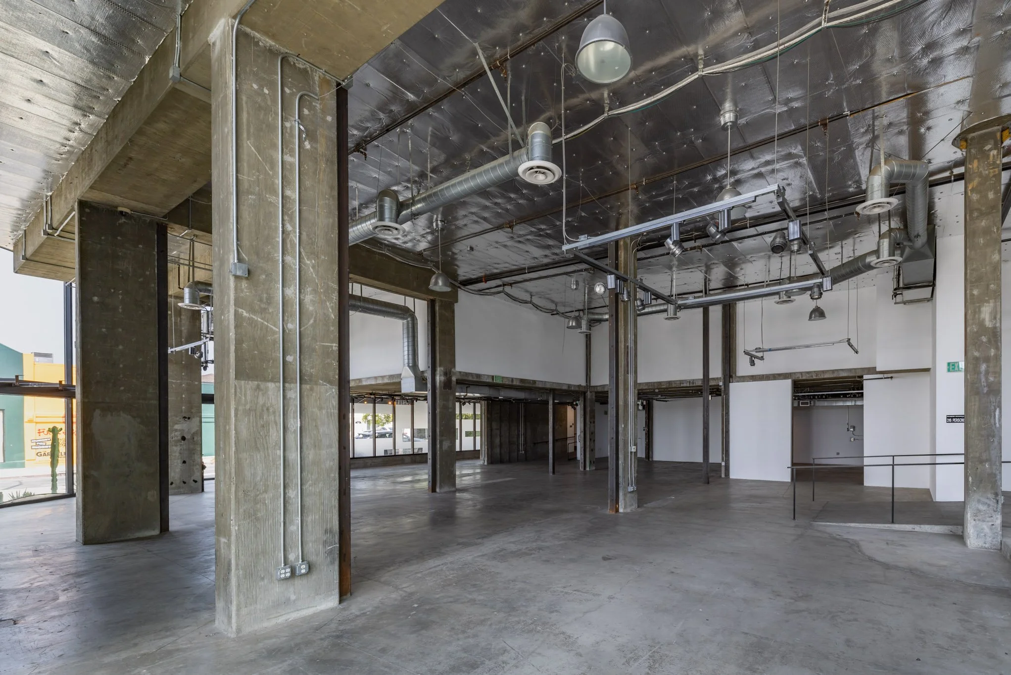 Empty commercial space with concrete floors and exposed ductwork and pipes on the ceiling, concrete support columns, and large glass windows on the left side.