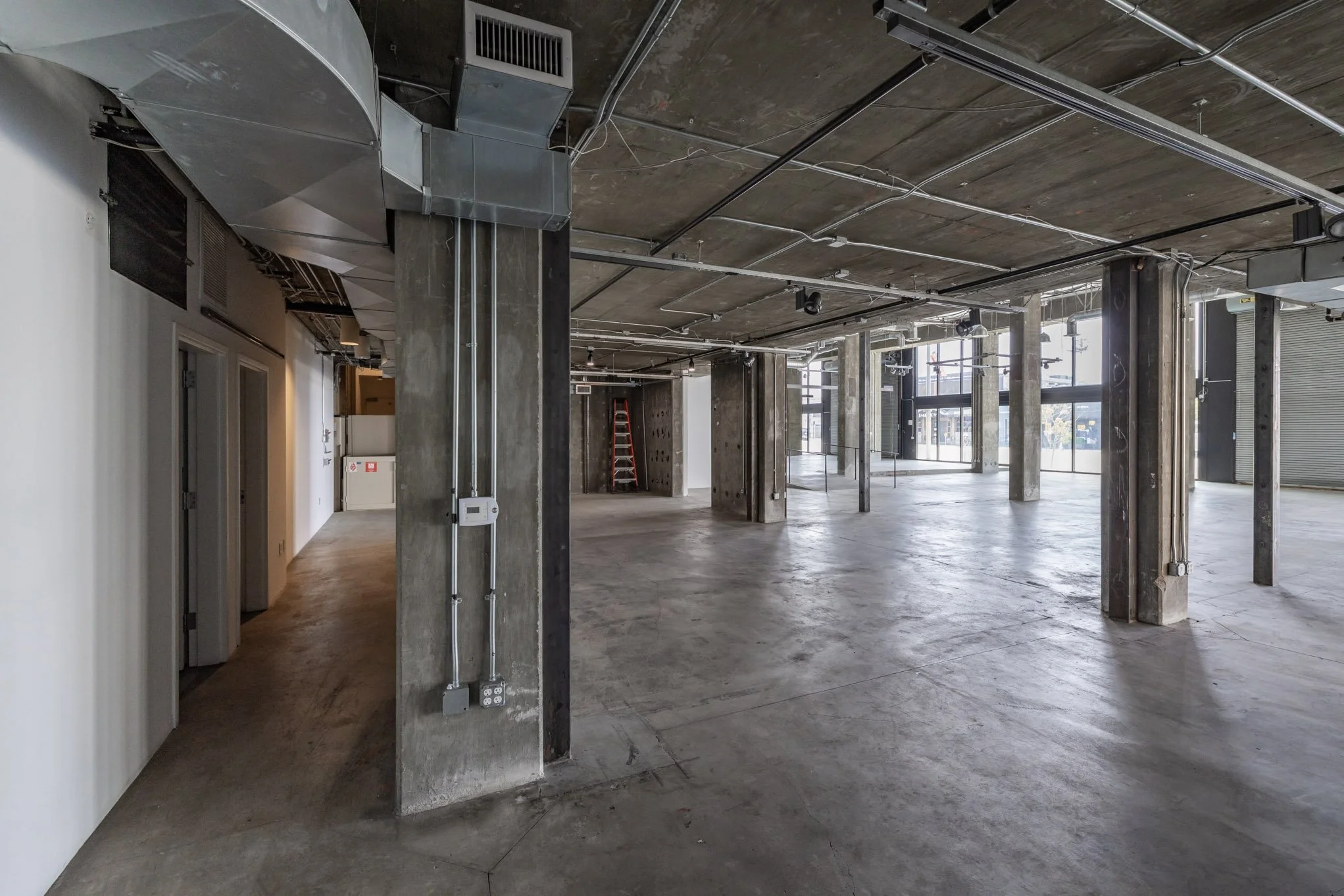 An unfinished commercial interior space with exposed concrete pillars, floors, and ceiling, electrical wiring, and large windows providing natural light.