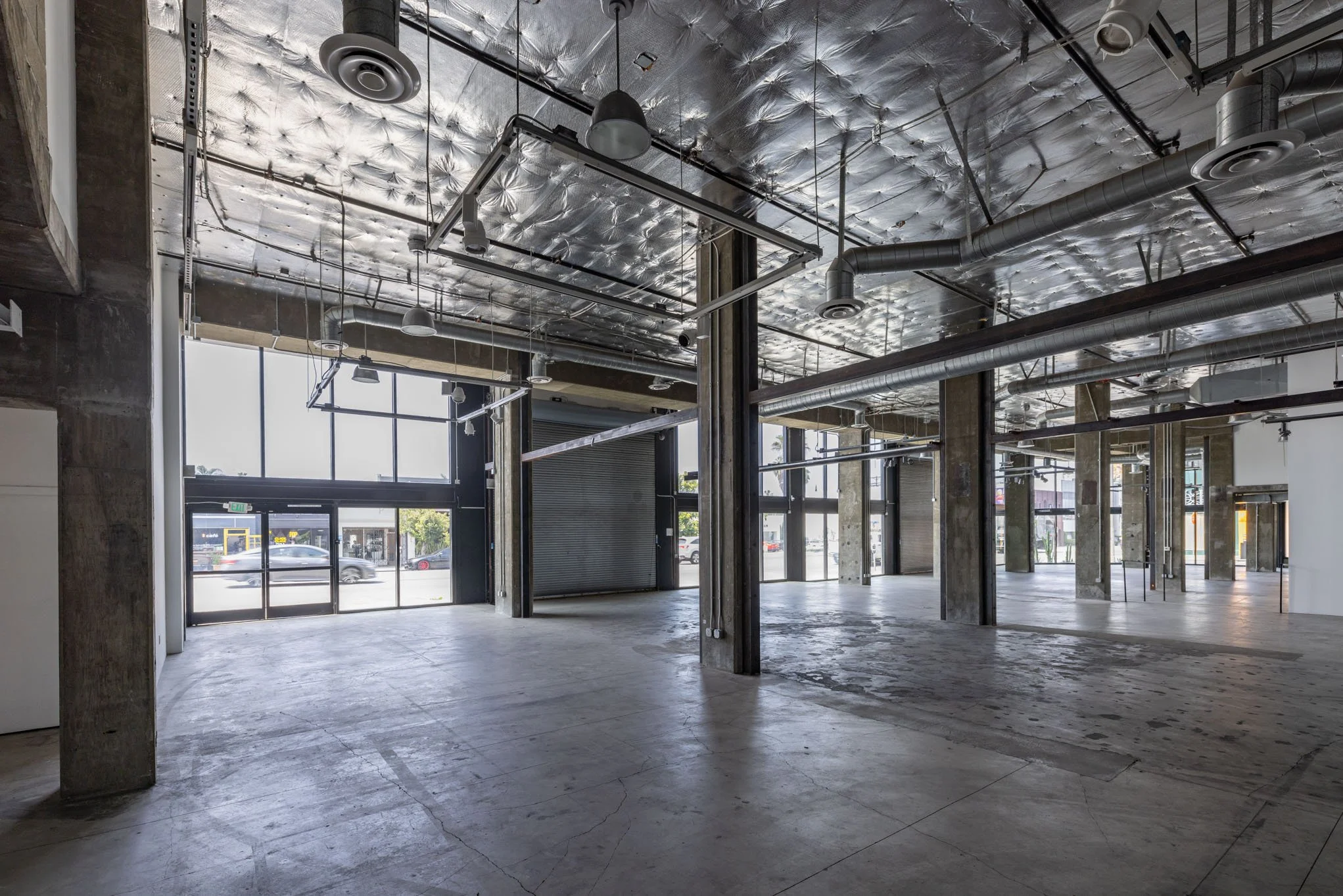 Empty commercial space with large windows, exposed ceiling pipes, and concrete pillars.