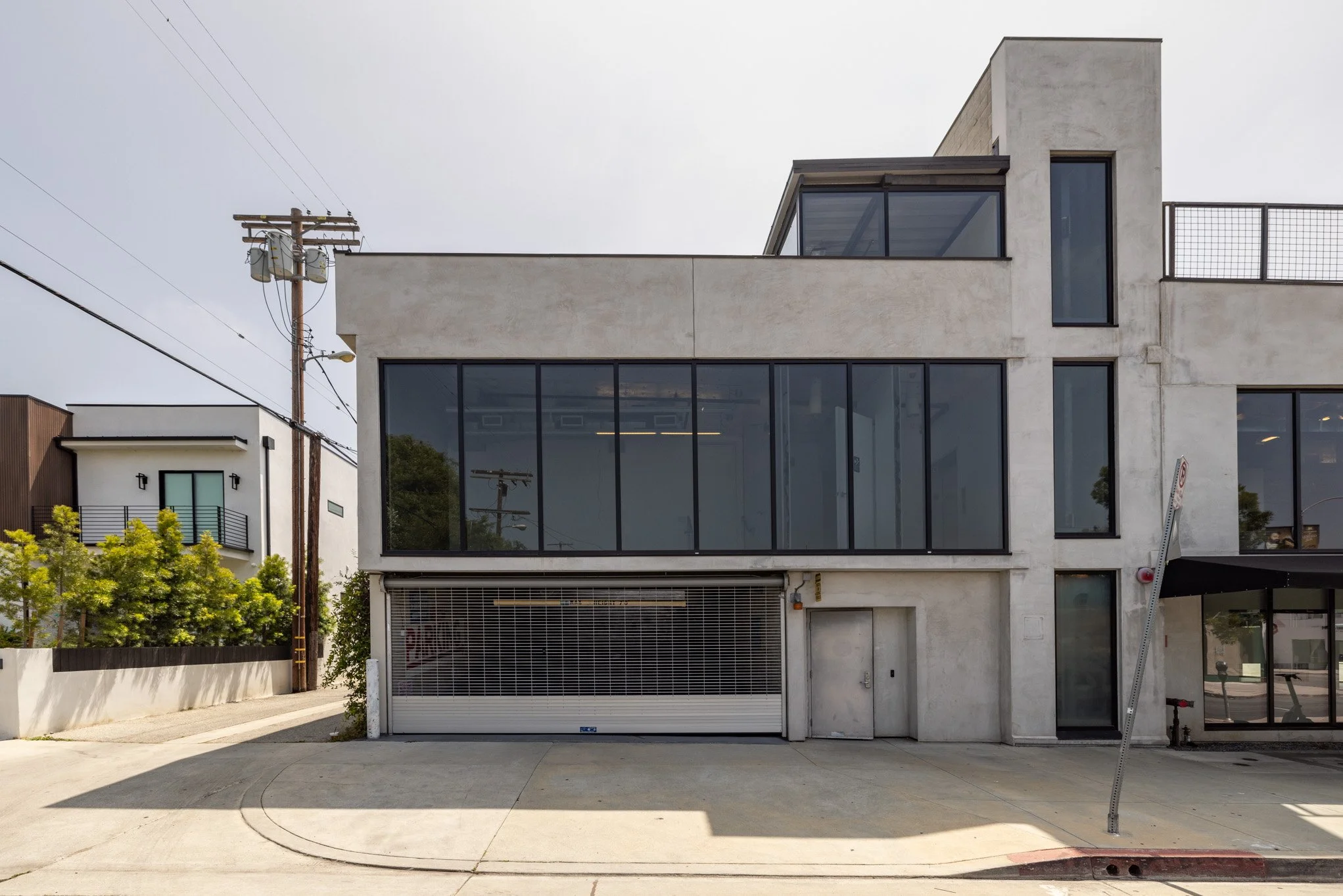 Modern multi-story building with large windows, white concrete exterior, and a garage door on the ground level. Power lines and other residential buildings are visible in the background, with cloudy sky overhead.
