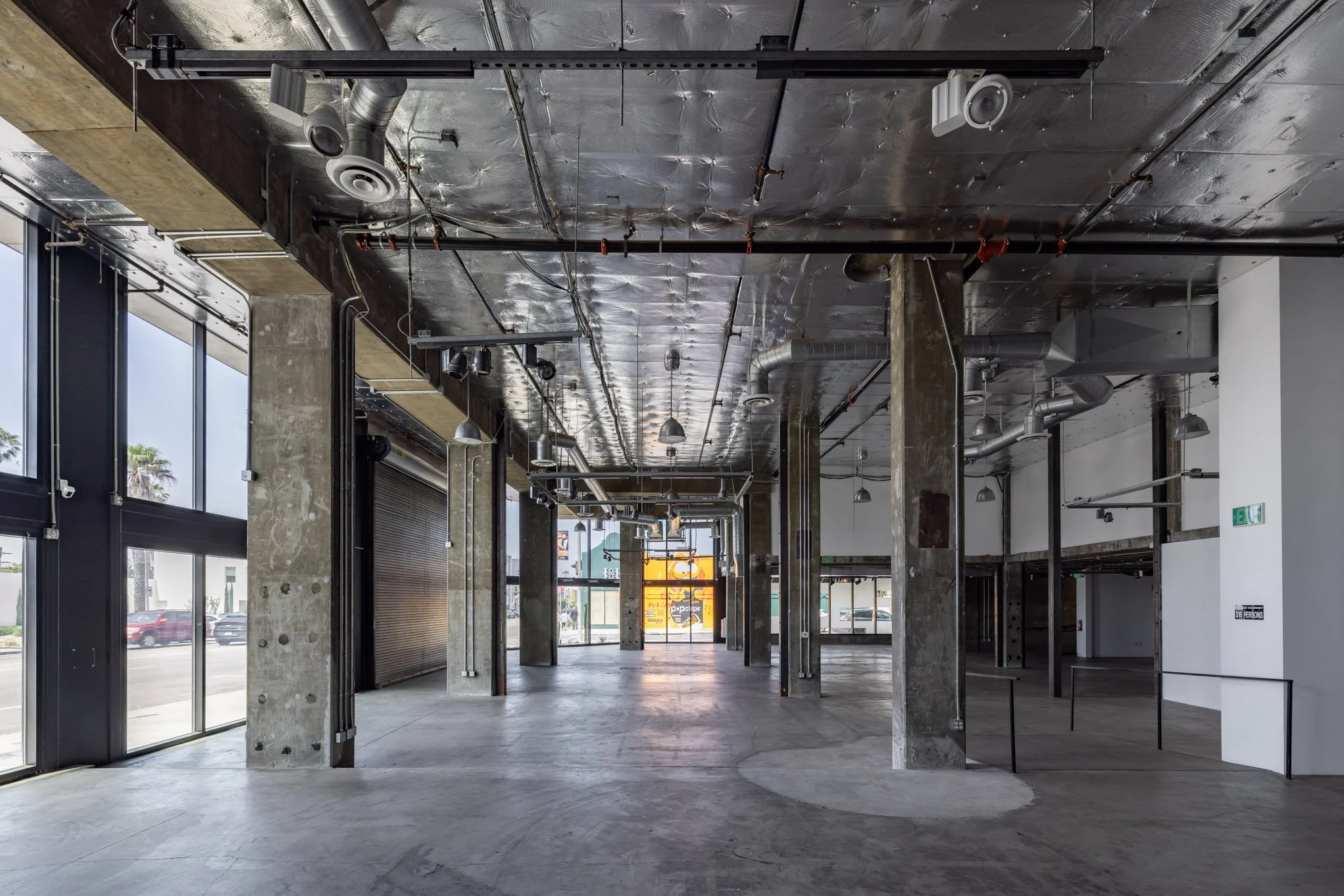 An empty commercial space with large glass windows, exposed concrete columns, and an unfinished ceiling with pipes and ductwork.