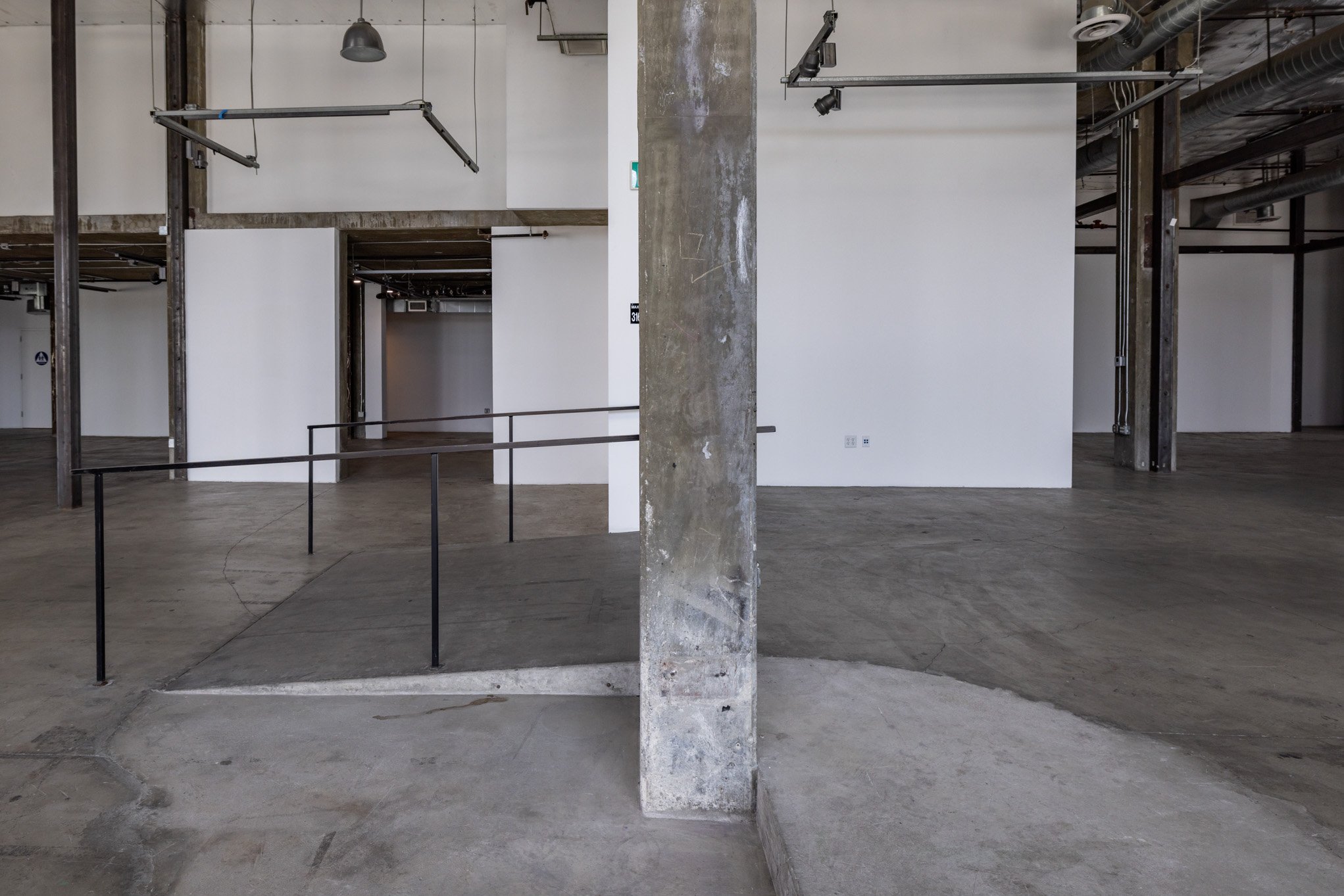 Empty industrial-style interior space with concrete floor, white walls, exposed beams and ductwork, a concrete pillar in the center, and a small ramp with a black railing.