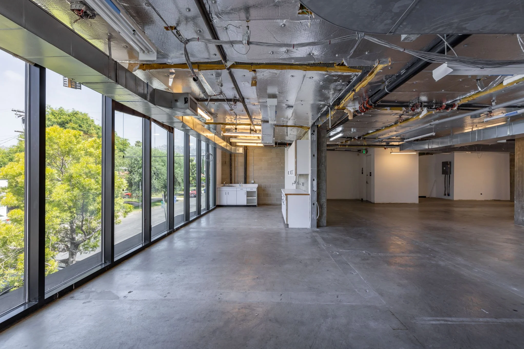 Empty commercial space under construction with large floor-to-ceiling windows, exposed ceiling with pipes, wires, and insulation, and unfinished concrete floor.