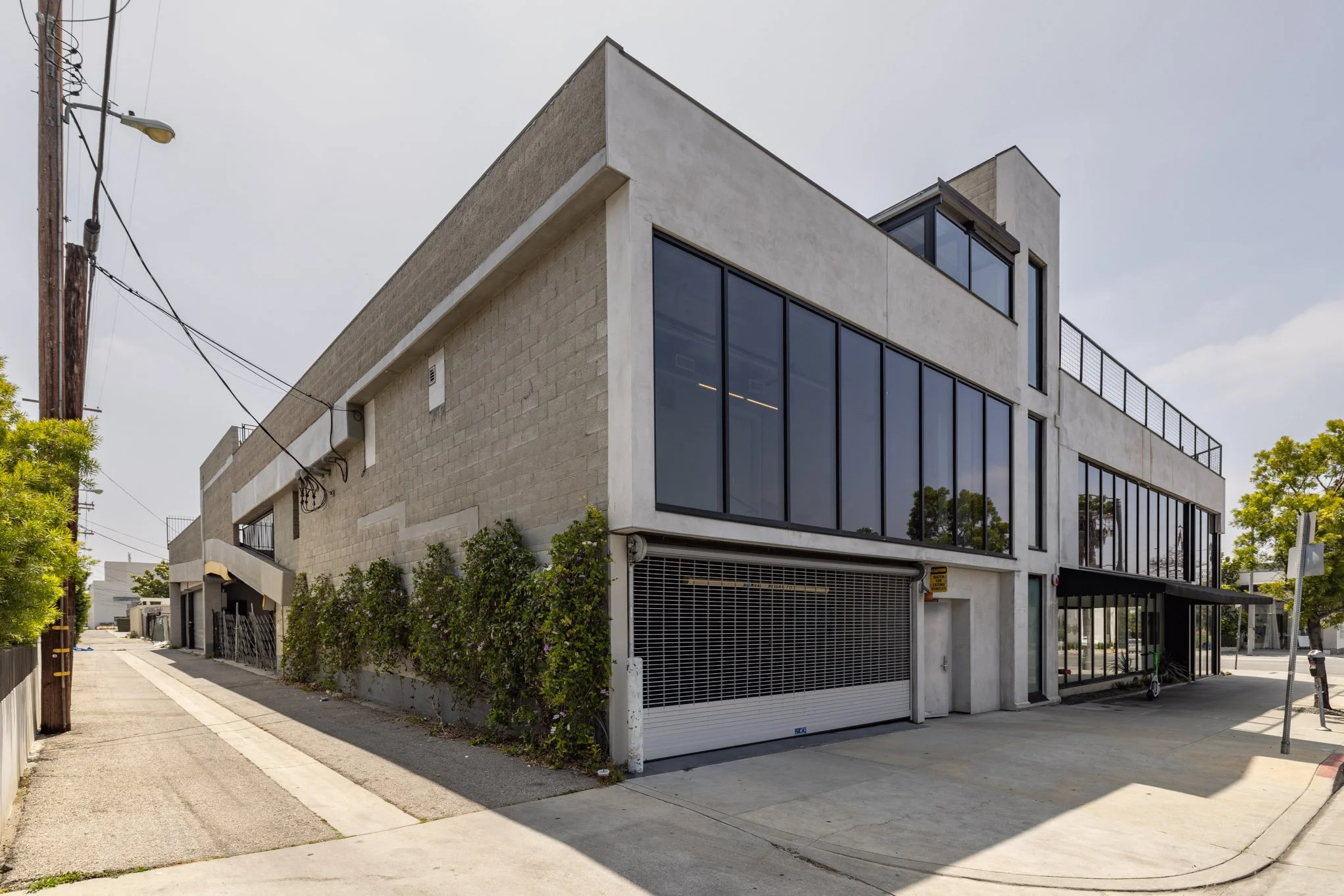 Modern multi-story building with large glass windows on a city street corner, surrounded by trees and concrete pavement.