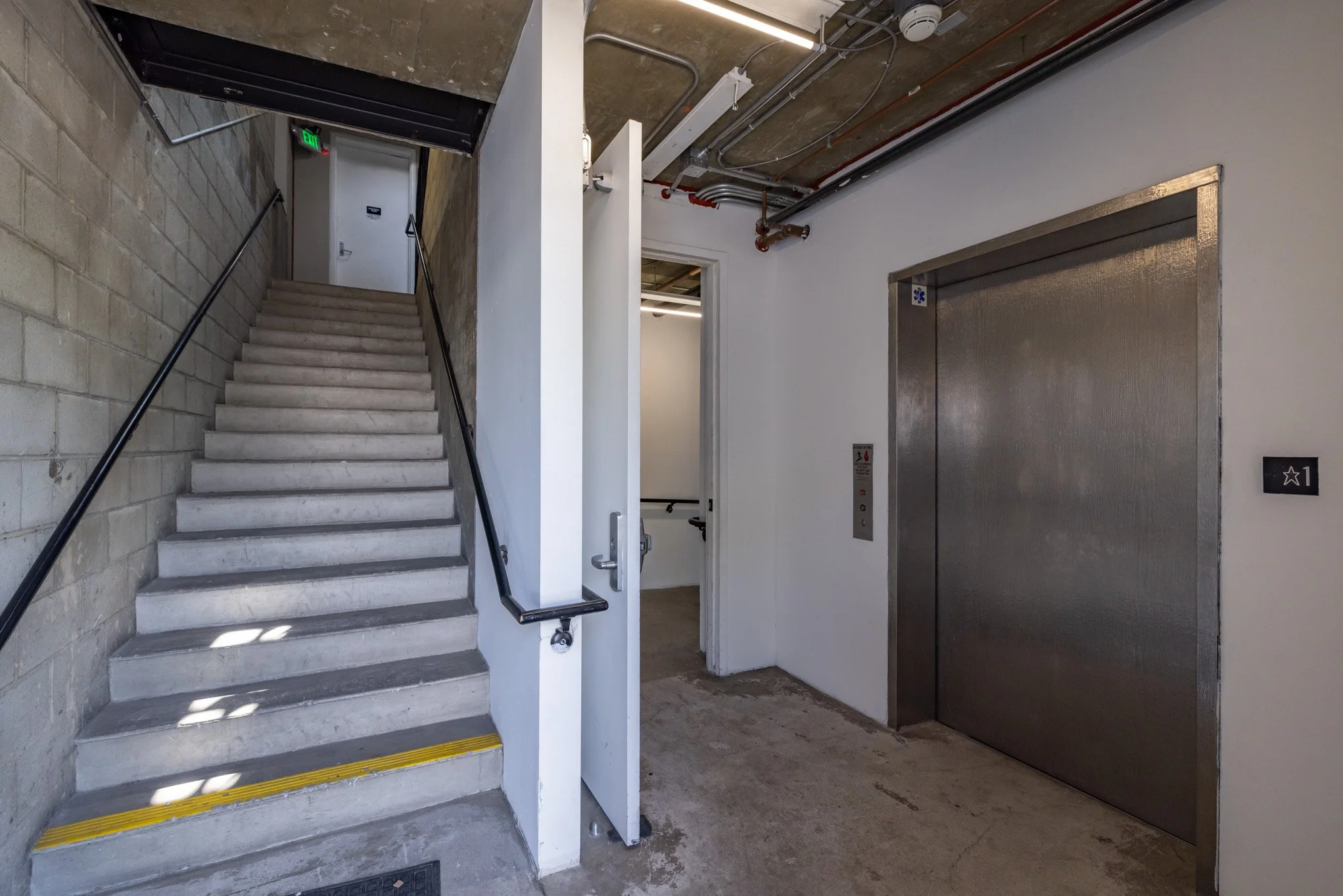 An indoor scene showing a staircase with black handrails, an elevator with metallic doors, and a room with an open door, in a building's stairwell or utility area.