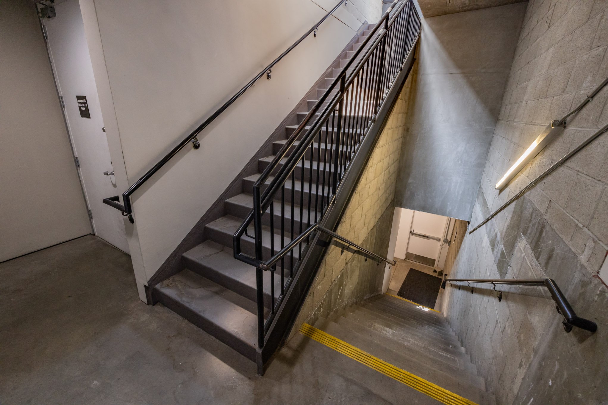 Indoor staircase with metal railings, yellow striped safety line on the stairs, beige brick wall on the right, and a gray wall with a door on the left. Fluorescent lighting on the wall.